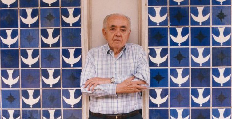 Athos Bulcão em pé, de braços cruzados, em frente ao painel azul de azulejos com pombas brancas e estrelas escuras, na Igreja Nossa Senhora de Fátima, em Brasília