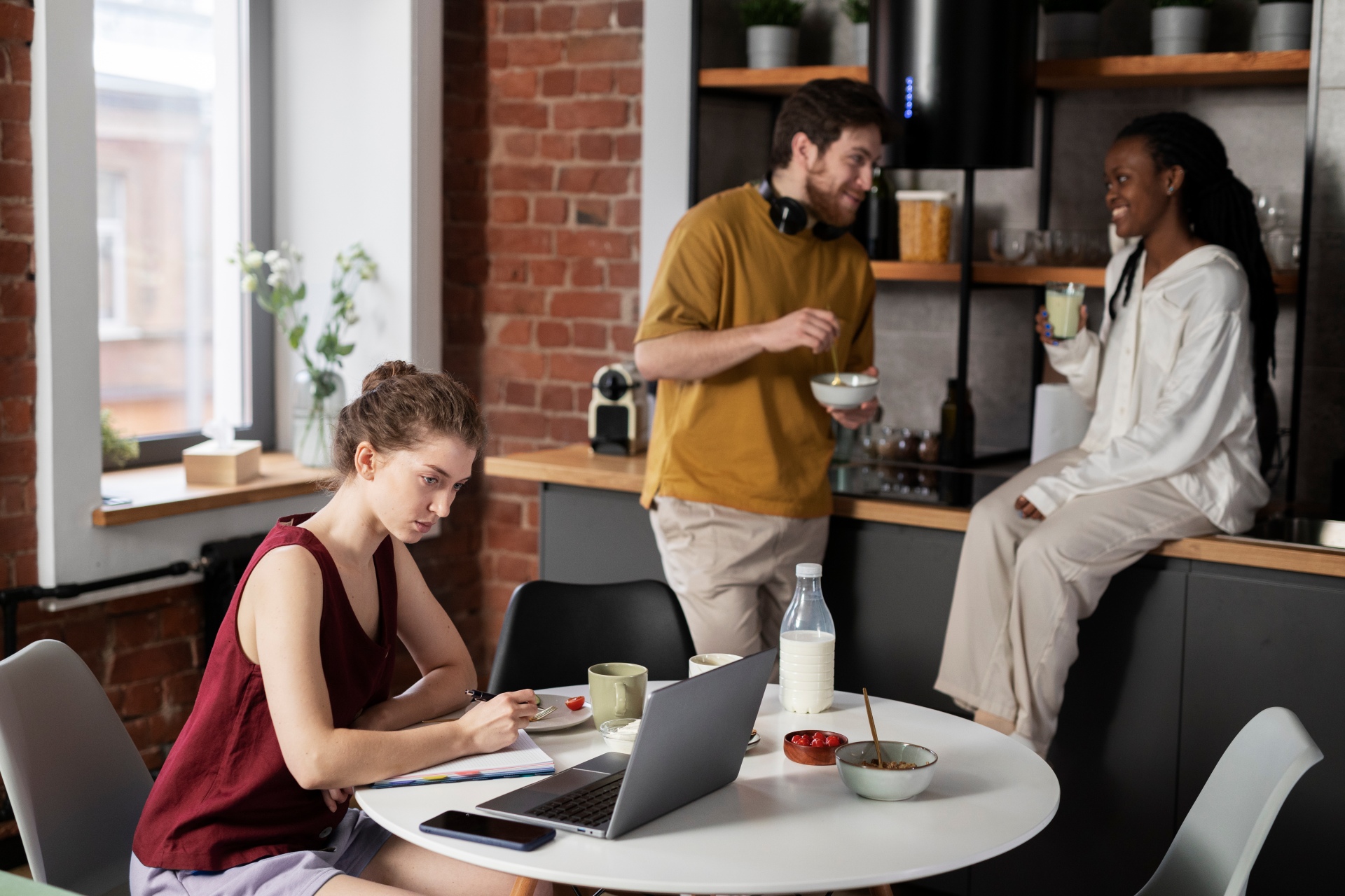 Colegas de quarto sorridentes em casa. Uma das mulheres está no notebook em uma mesa. Um homem e uma mulher conversam ao fundo na cozinha.
