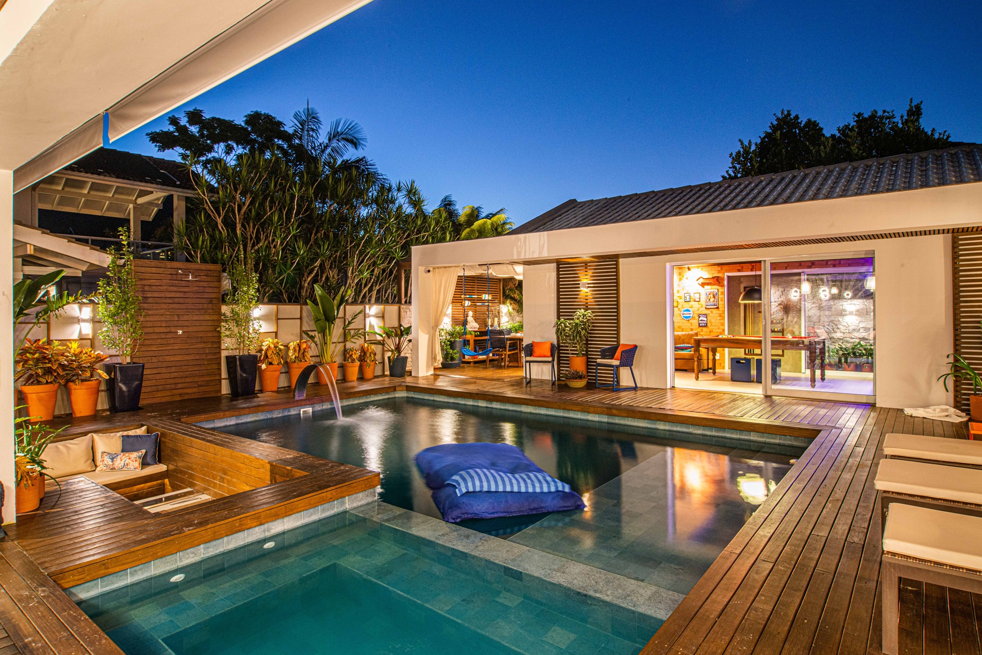 Fachada interna de uma casa com piscina, detalhes e chão de madeira, com muitos pontos de luz e plantas.