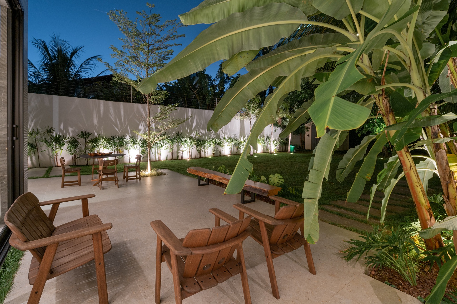 Área externa de uma casa revestida com porcelanato, cadeiras de madeira, plantas e iluminação.
