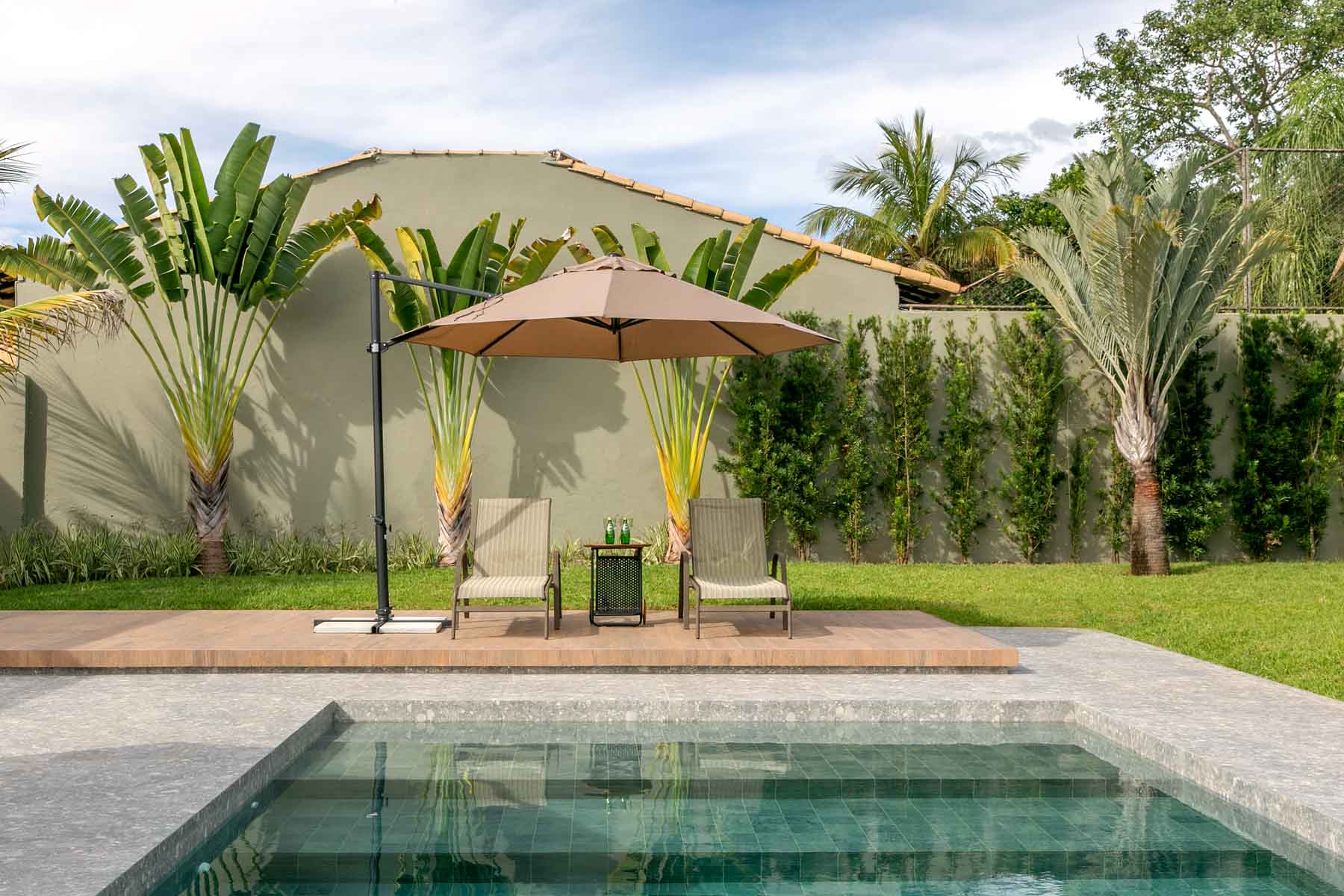 Área externa de uma casa, jardim com piscina e dois cadeiras de praia embaixo de um guarda-sol.