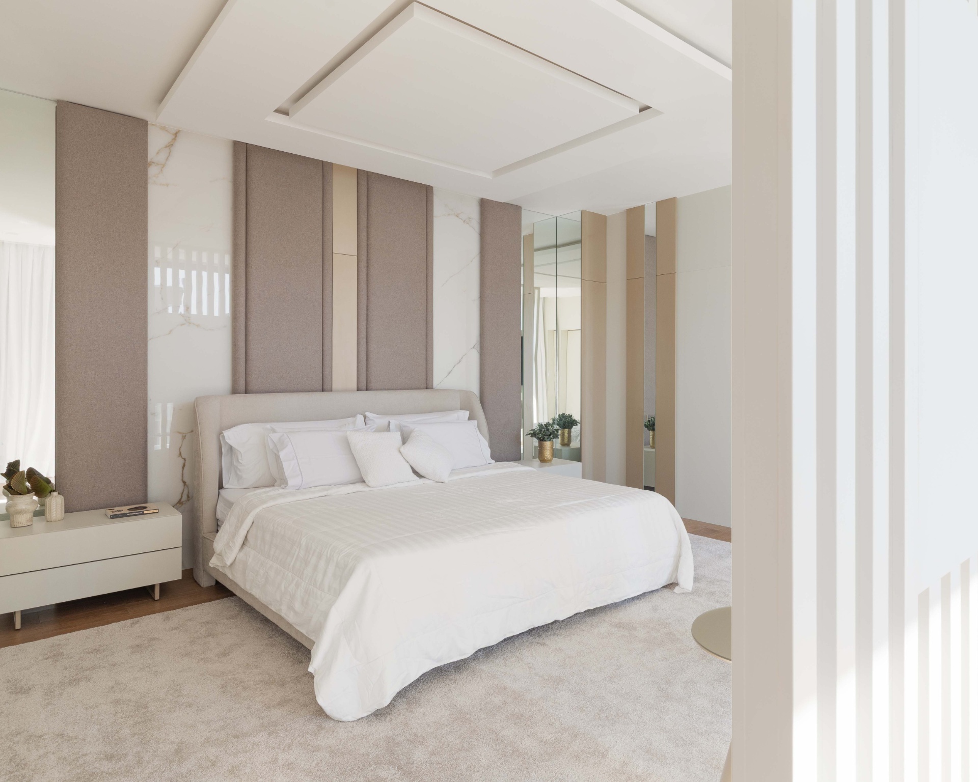 Quarto de casal em tons neutros, com cama e tapete felpudo. Na parede, há um espelho grande
