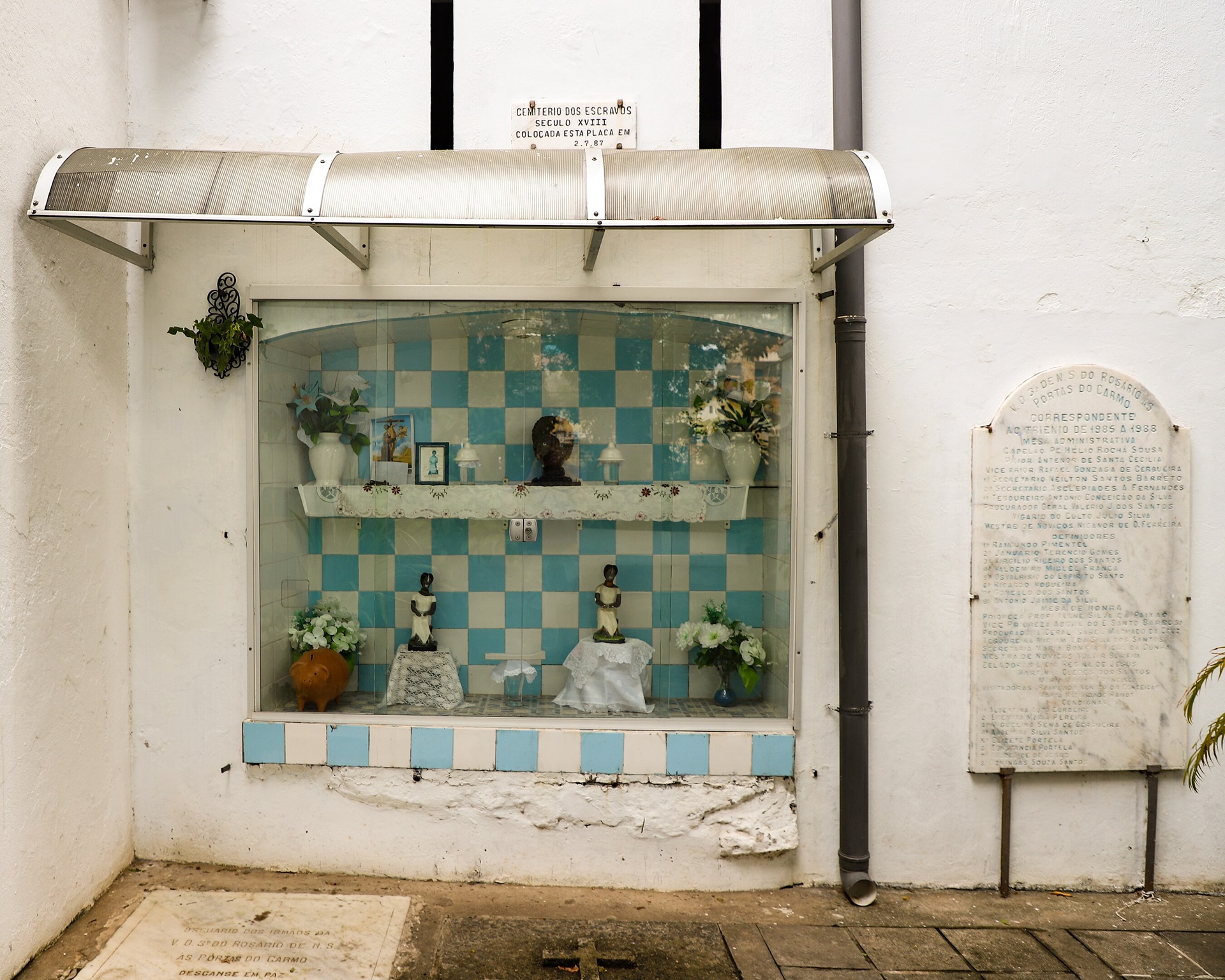 igrejas pelo mundo Memorial do Cemitério dos Escravizados, nos fundos da Igreja do Rosário dos Pretos, em Salvador, com vitrine protegida exibindo imagens devocionais e painel azul e branco ao fundo.