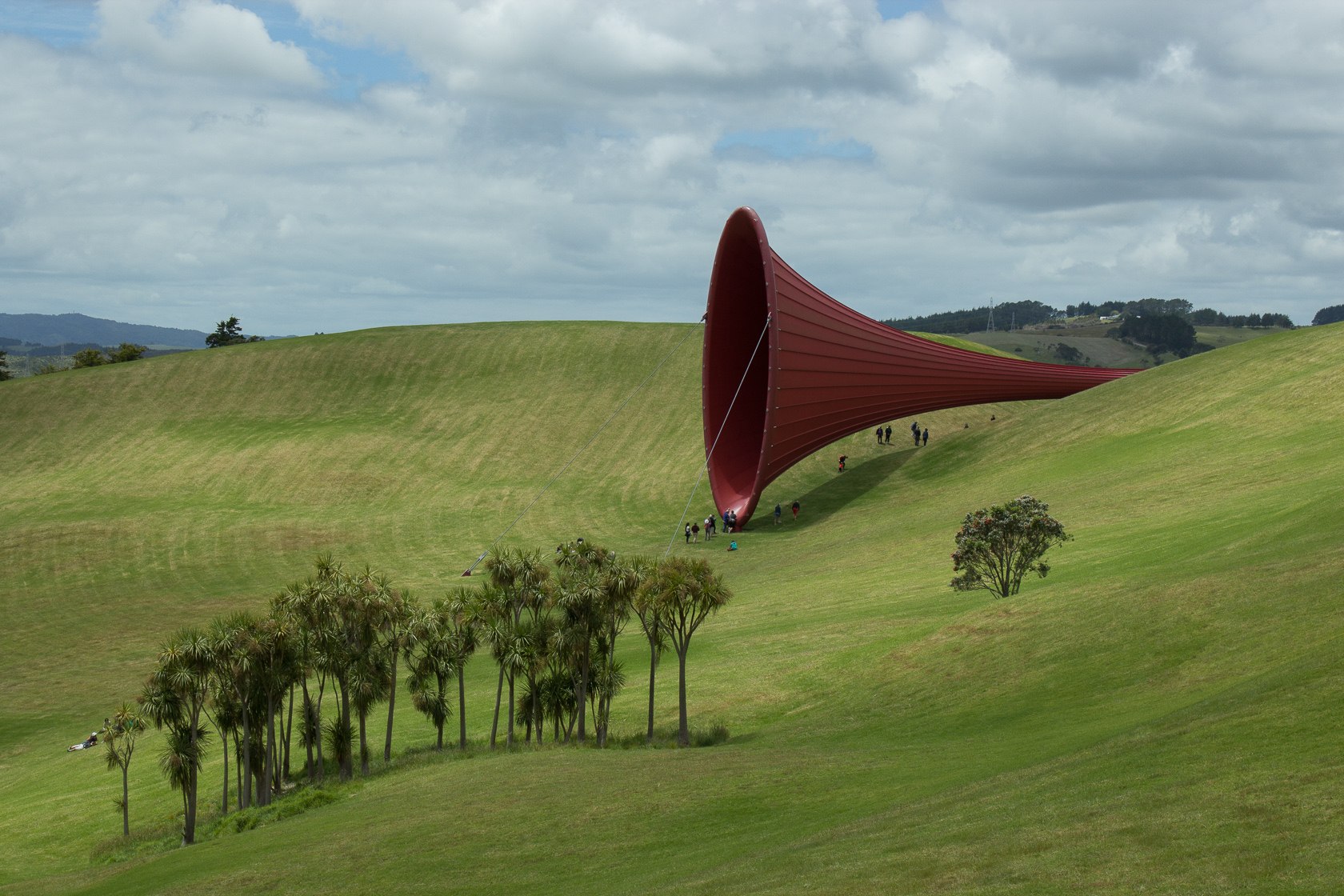 Em meio a um campo, visitantes apreciam uma escultura vermelha gigante em formato de corneta