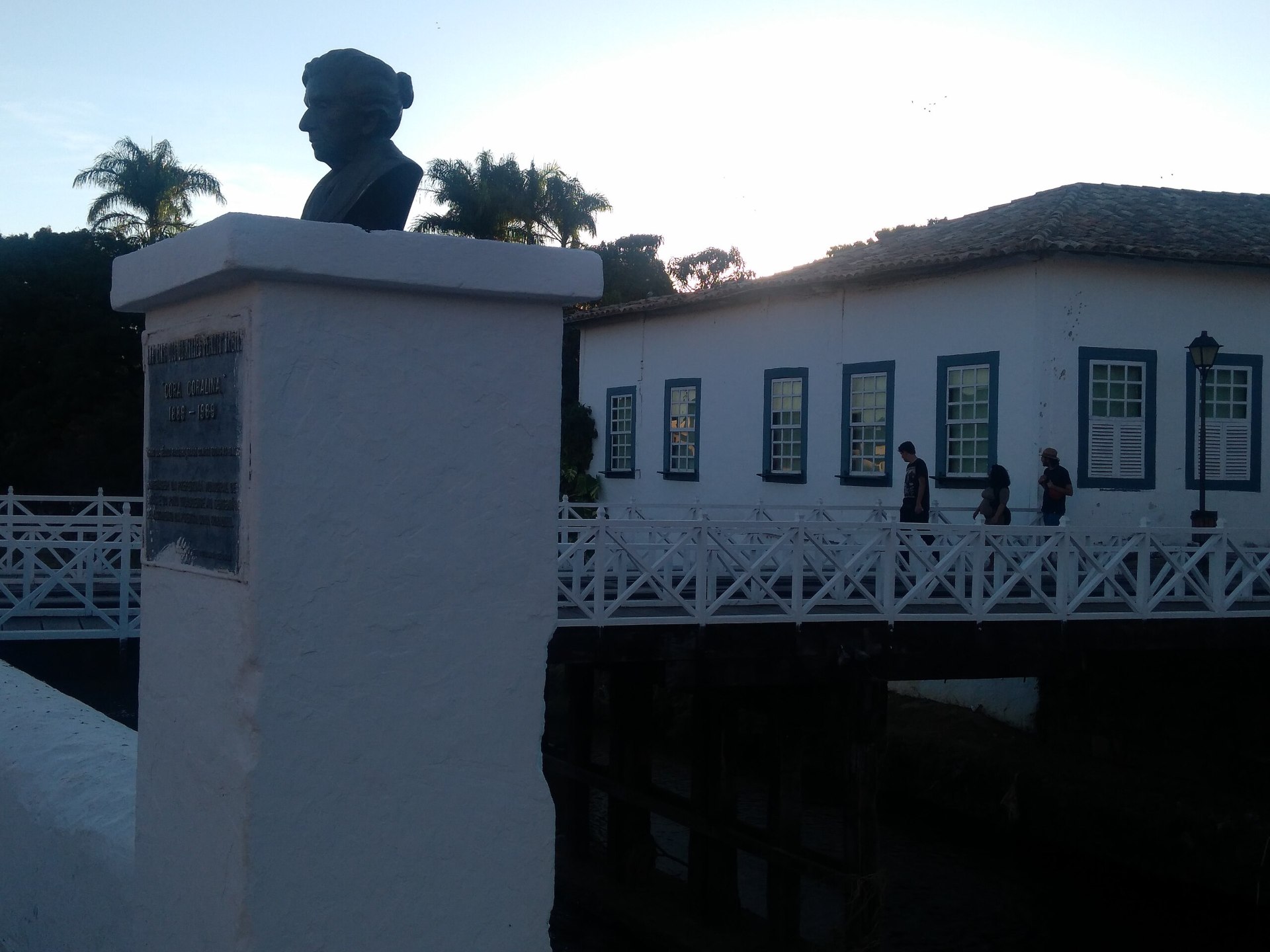 Busto de Cora Coralina sobre pilar de concreto. Ao fundo, é possível observar uma ponte, com pessoas passando, e parte de uma casa de estilo colonial, com diversas janelas