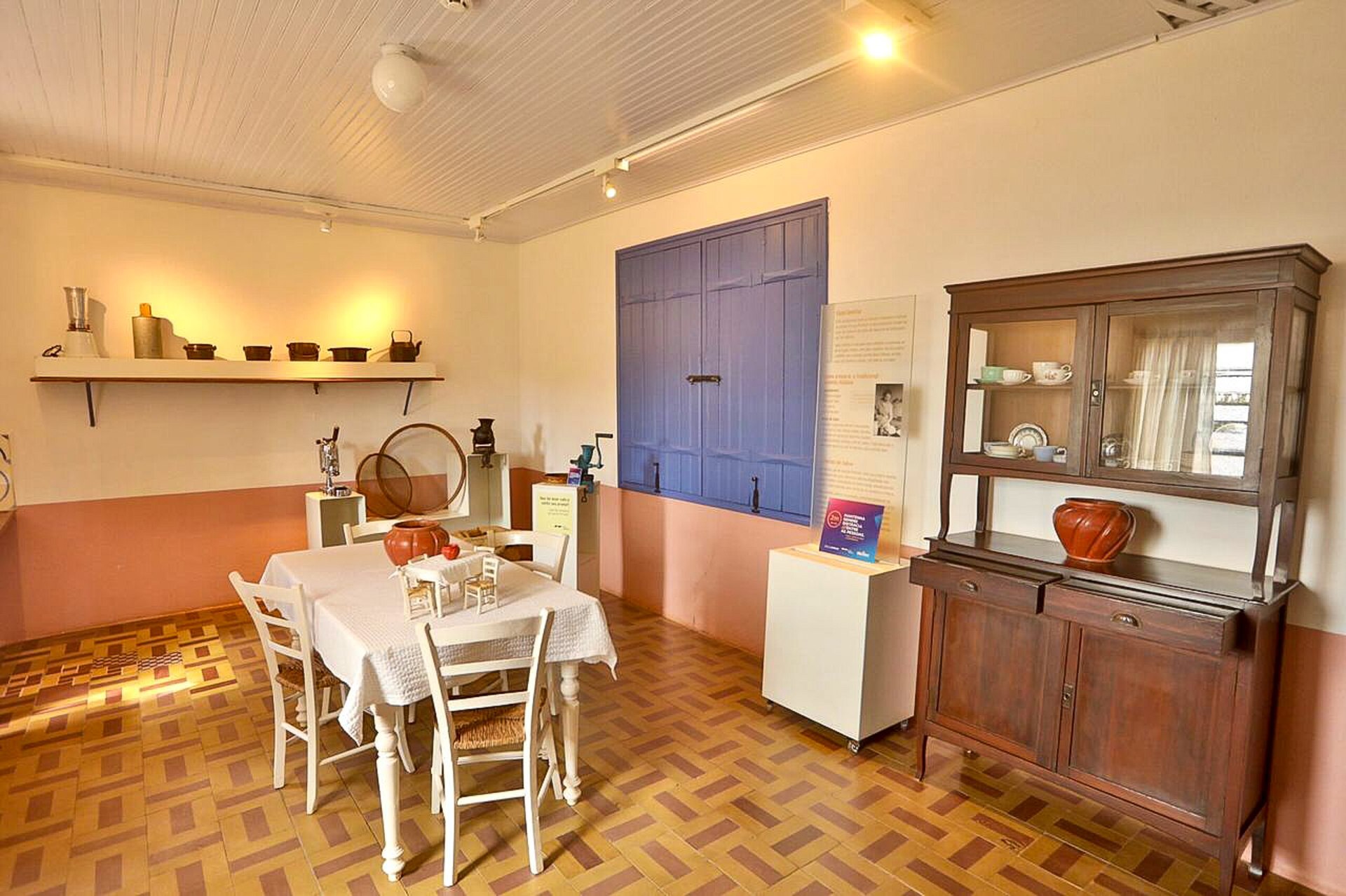 Cozinha do Museu Casa de Portinari, onde há uma mesa branca ao centro, uma janela azul e uma cristaleira encostada em uma das paredes