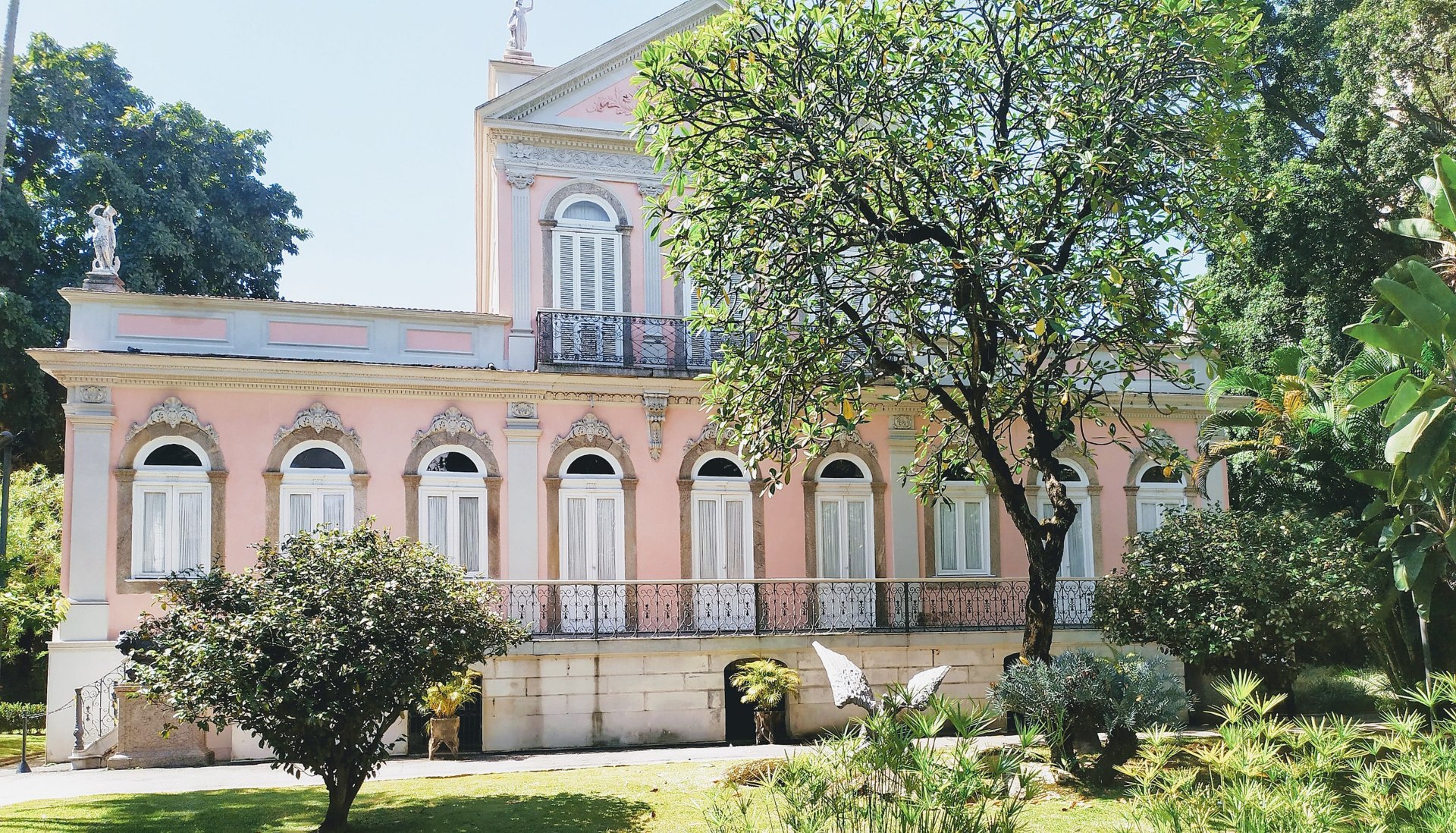 Fachada rosa-claro de casa de dois andares, em arquitetura neoclássica, com um jardim em frente