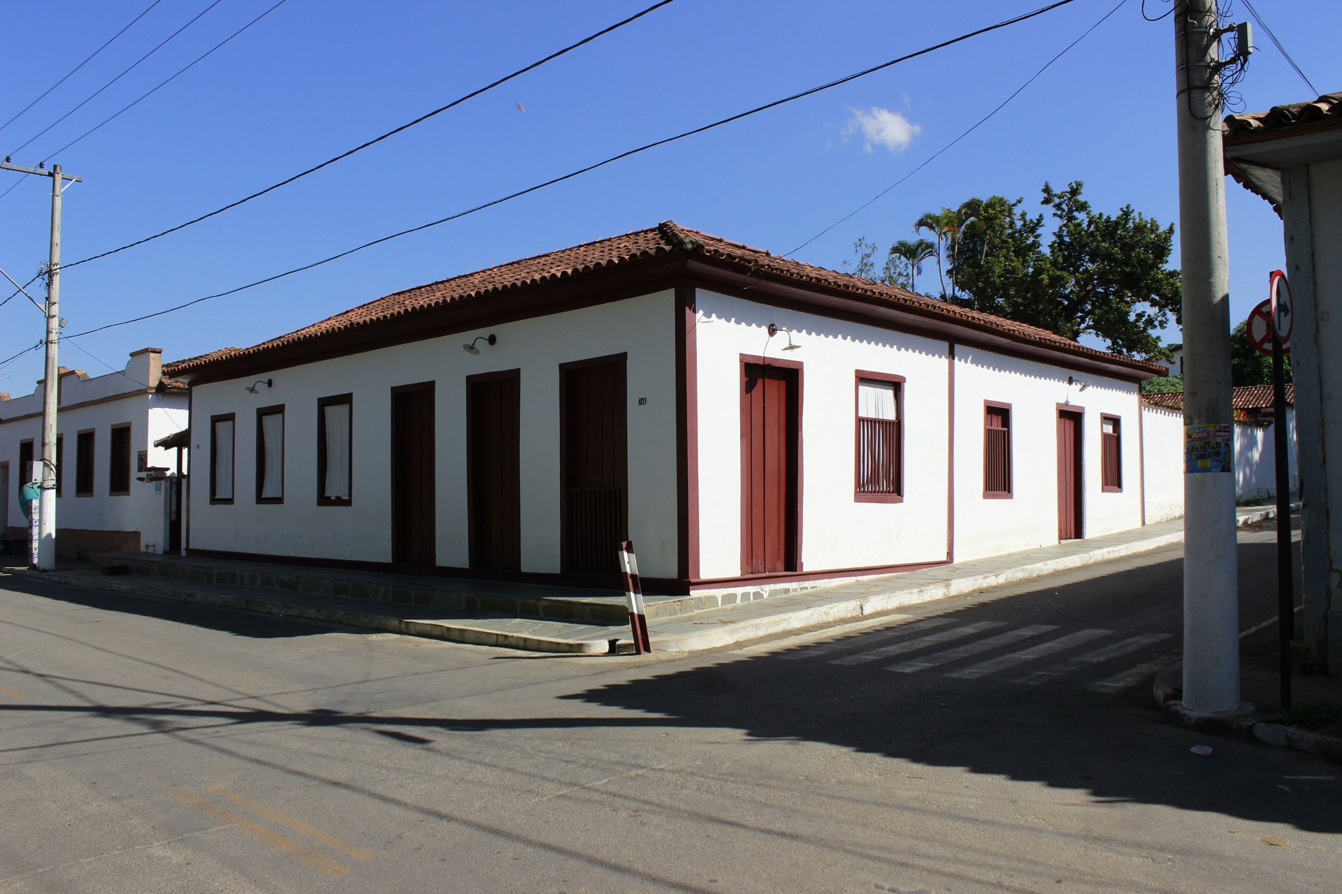 Grande casa em estilo colonial, observada a partir de uma esquina. Possui paredes brancas e portas e janelas de madeira na cor marrom
