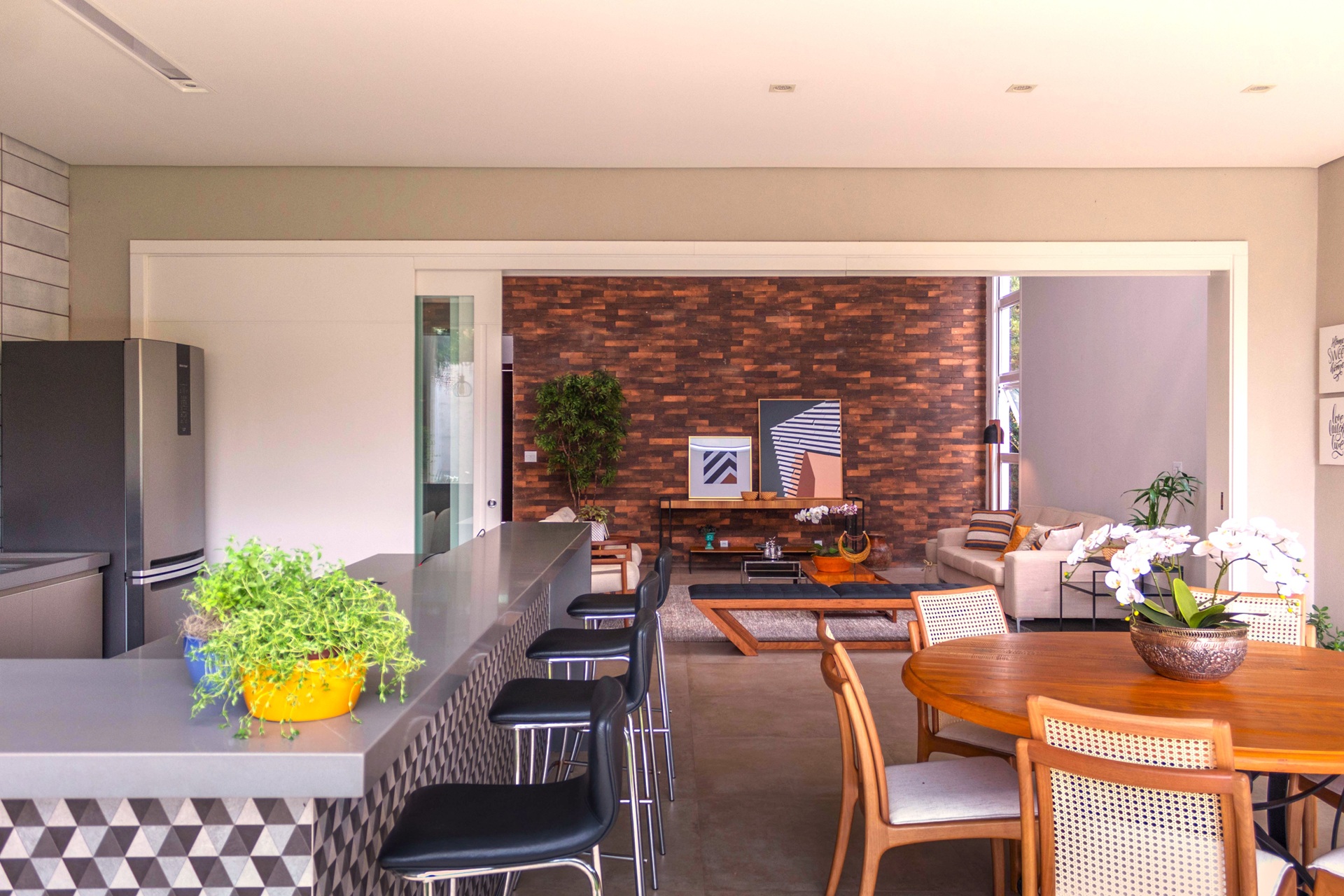 orquídea Ambiente integrado com cozinha, sala de jantar e sala de estar; destaque para orquídeas brancas sobre a mesa redonda de madeira e no aparador da sala, com parede de tijolos e decoração contemporânea ao fundo.