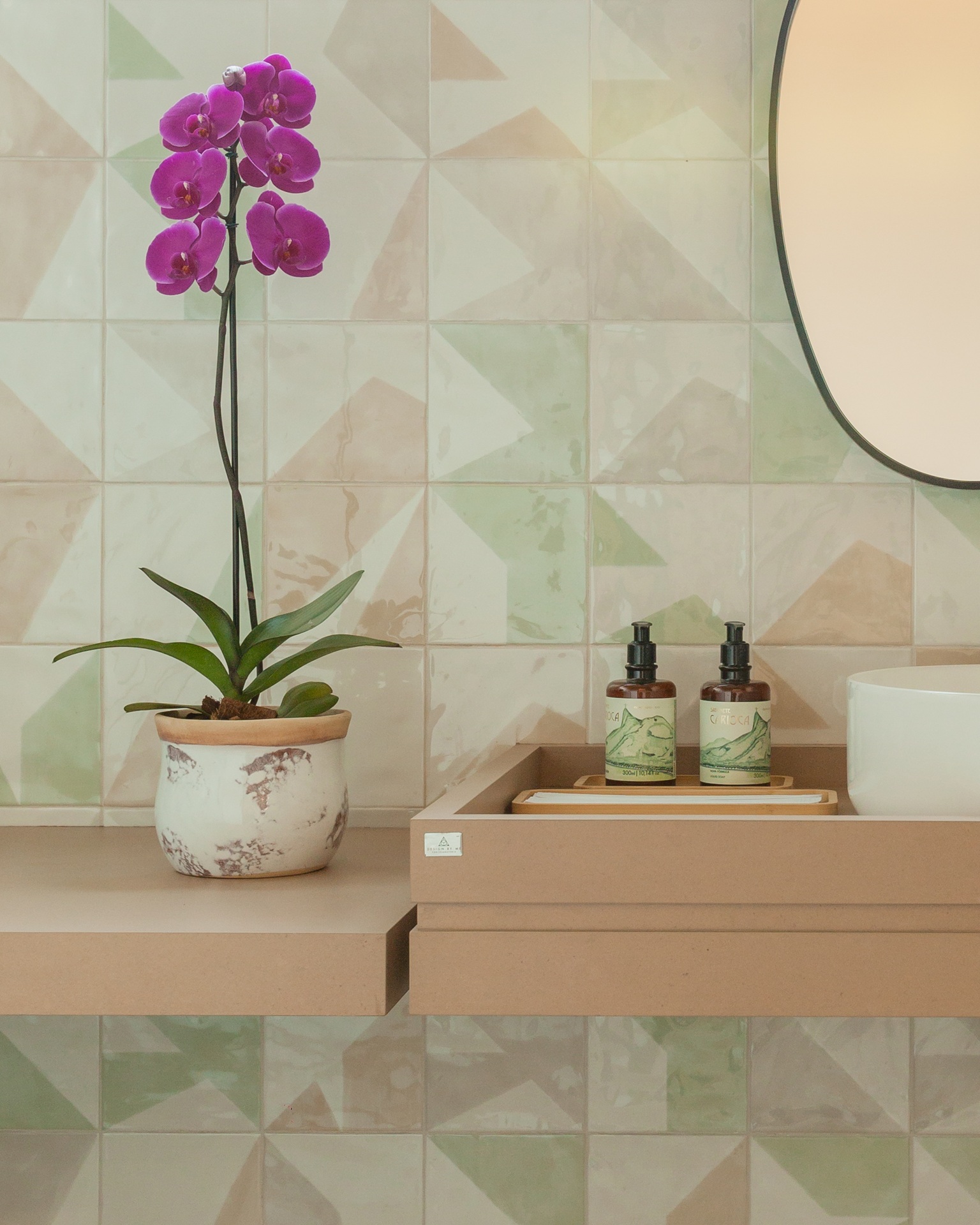 orquídea Orquídea rosa vibrante em vaso marmorizado sobre bancada de banheiro com revestimento geométrico em tons pastel, ao lado de bandeja com sabonetes líquidos e espelho oval.