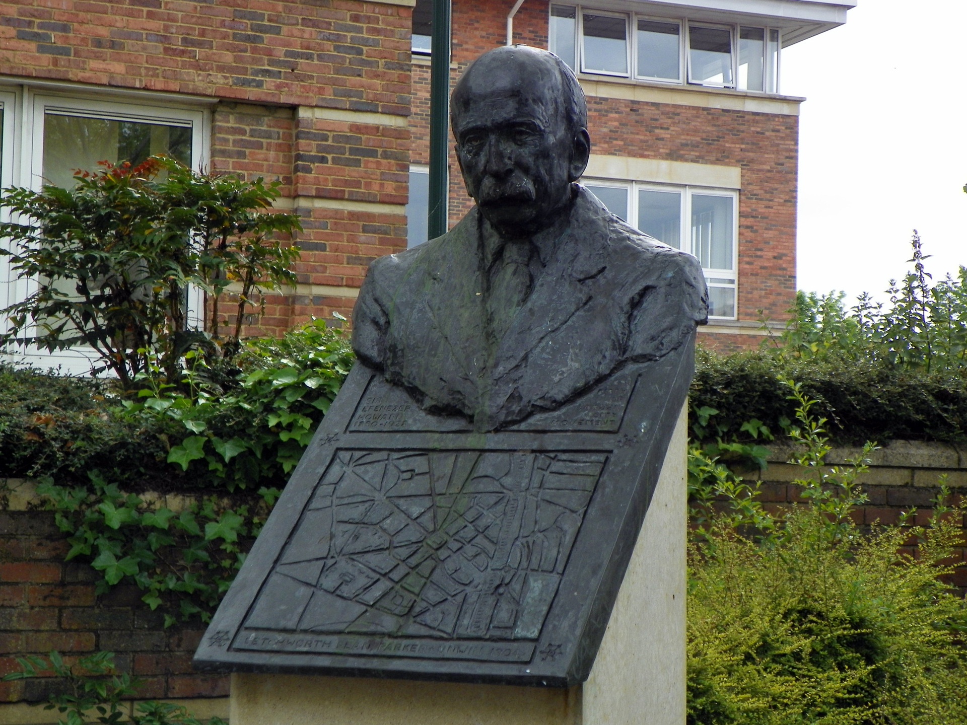 cidades-jardim Busto de bronze de Ebenezer Howard, localizado em Letchworth Garden City, Inglaterra. A escultura repousa sobre um pedestal inclinado, que exibe em relevo um mapa do plano original da cidade-jardim, cercada por vegetação e com edifícios de tijolos aparentes ao fundo.