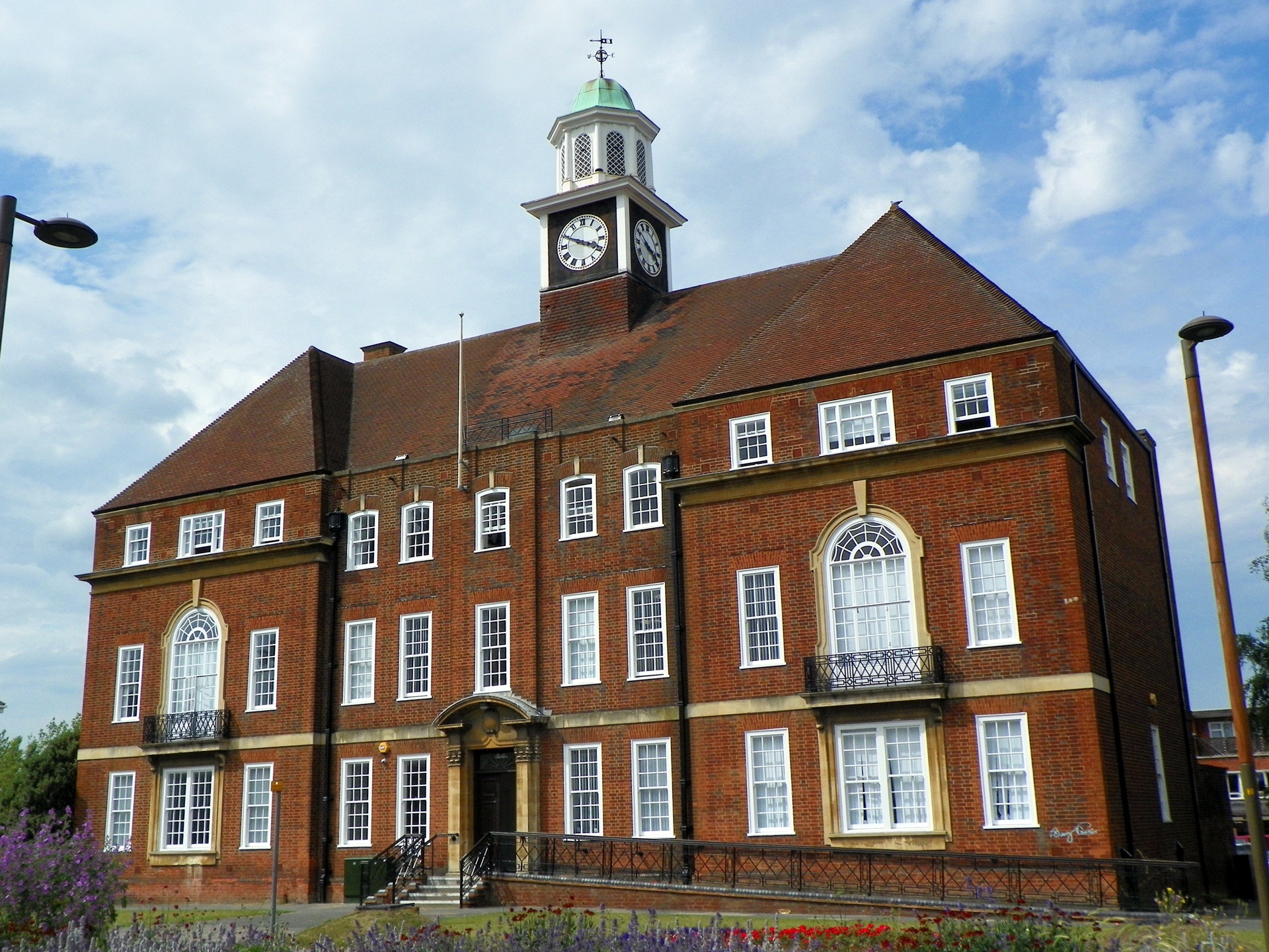 Fachada da prefeitura de Letchworth Garden City, Inglaterra. O edifício de tijolos vermelhos possui estilo georgiano, com janelas simétricas, sacadas em ferro e um relógio sobre a torre central. À frente, um canteiro florido emoldura o prédio, refletindo o cuidado estético e paisagístico típico das cidades-jardim.