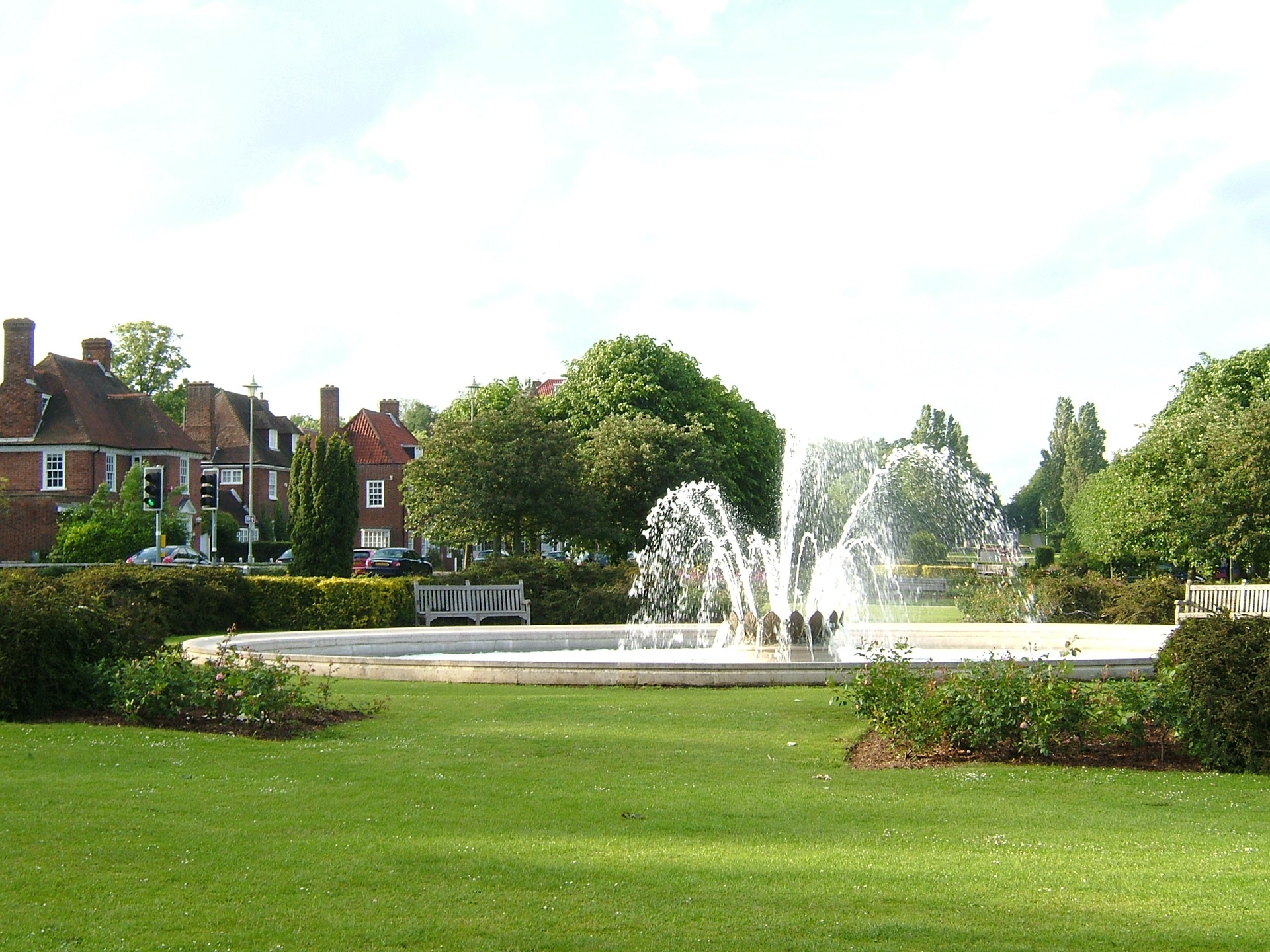 cidades jardim Vista da Fonte da Coroação, localizada na Parkway, em Welwyn Garden City, Inglaterra. Cercada por gramados bem cuidados, árvores e bancos de madeira, a fonte centraliza o espaço público e reforça a vocação paisagística da cidade-jardim, com casas de tijolos vermelhos ao fundo e um ambiente que privilegia o convívio e a harmonia com o verde.