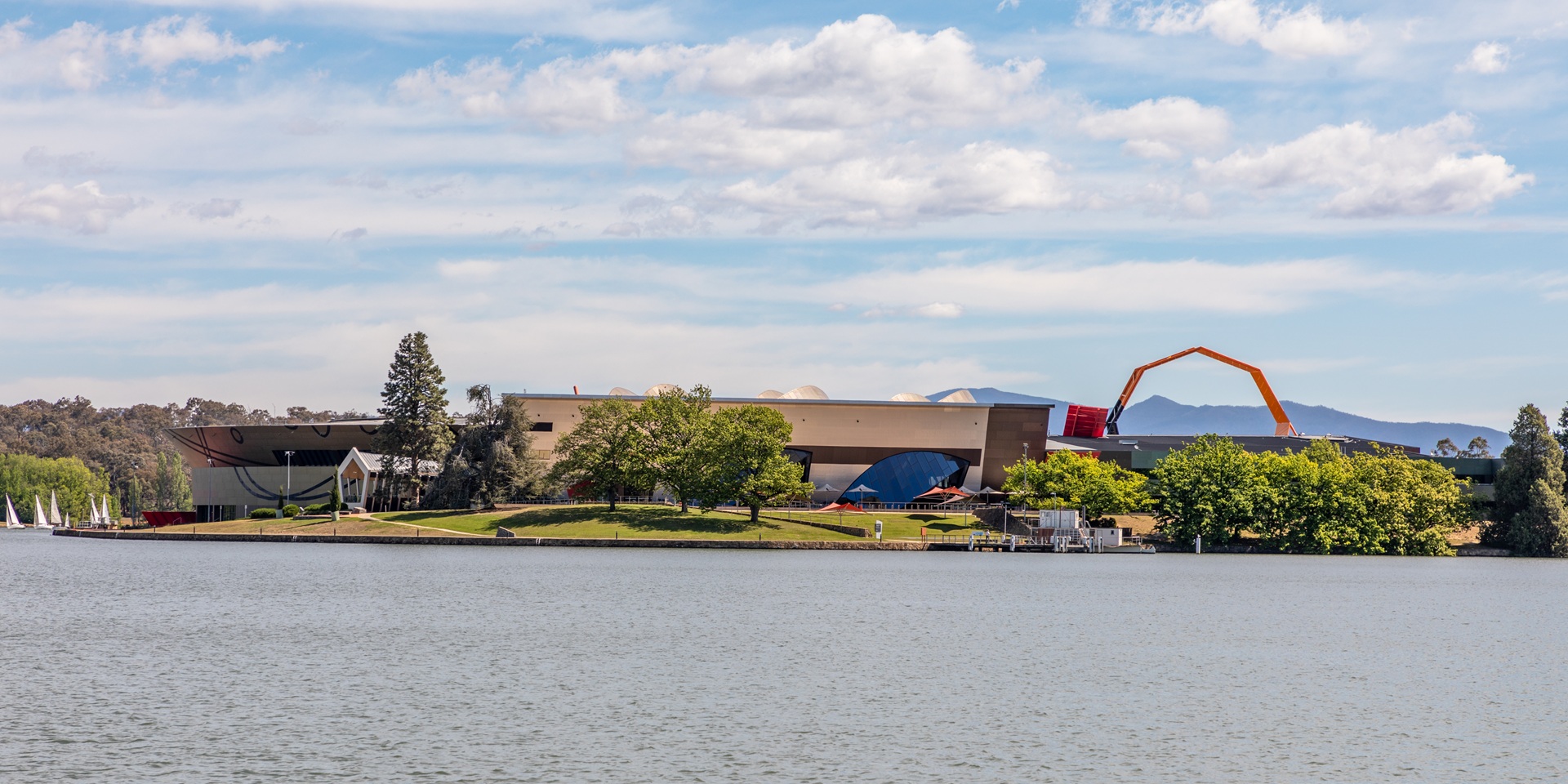 cidade jardim Vista do National Museum of Australia em Canberra, com sua arquitetura contemporânea marcante, à beira do lago Burley Griffin. A paisagem evidencia a integração entre edificações culturais, espaços verdes e espelhos d’água — característica essencial do conceito de cidade-jardim adotado no planejamento da capital australiana. Ao fundo, é possível observar as montanhas e a vegetação nativa que envolvem a cidade.