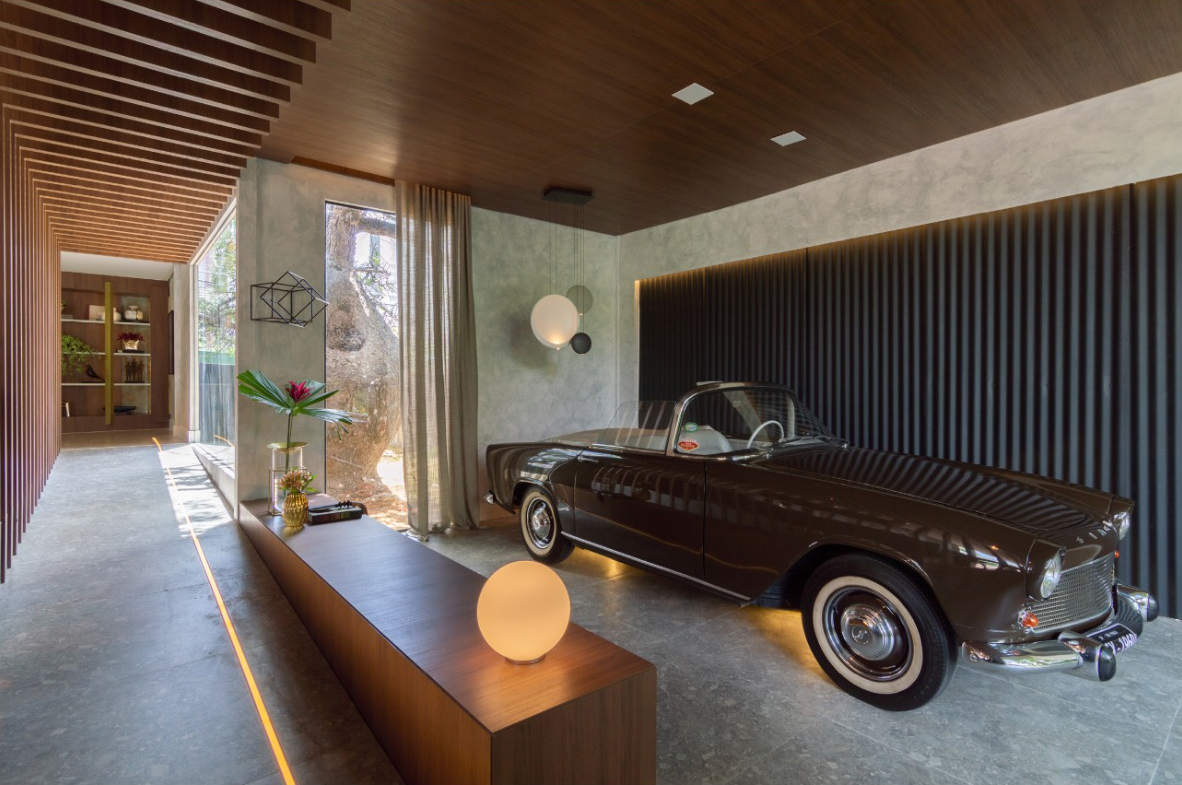 Ambiente que valoriza a decoração da garagem de estar com estética refinada: piso em porcelanato, painéis ripados de madeira no teto e parede, iluminação indireta e mobiliário minimalista. O carro antigo compõe o espaço como elemento decorativo, destacando o equilíbrio entre funcionalidade e elegância