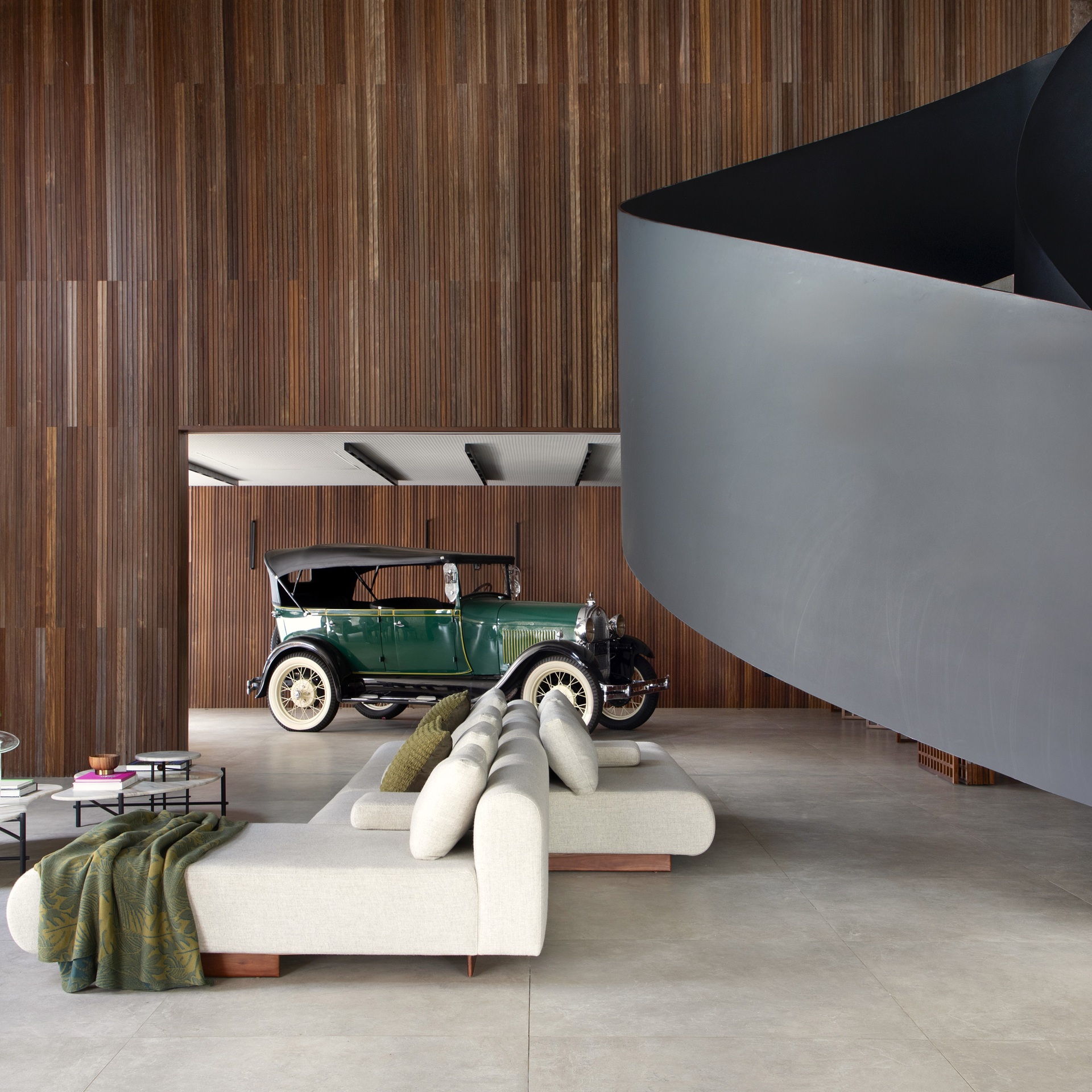 Garagem de estar elegante e minimalista, onde o carro antigo se destaca como peça de design integrada ao ambiente de convívio. Com sofá moderno, piso em porcelanato e painéis ripados de madeira, o espaço valoriza a multifuncionalidade e o aproveitamento inteligente da área, além de contribuir para a estética e a valorização do imóvel