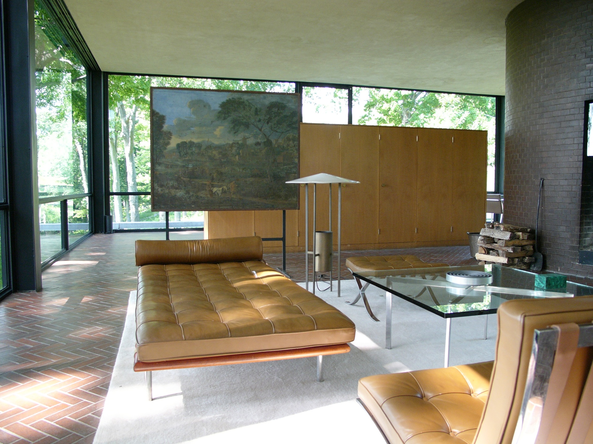 Interior da Glass House, com móveis de couro, mesa de vidro, lareira de tijolos e vista para a vegetação através das paredes envidraçadas