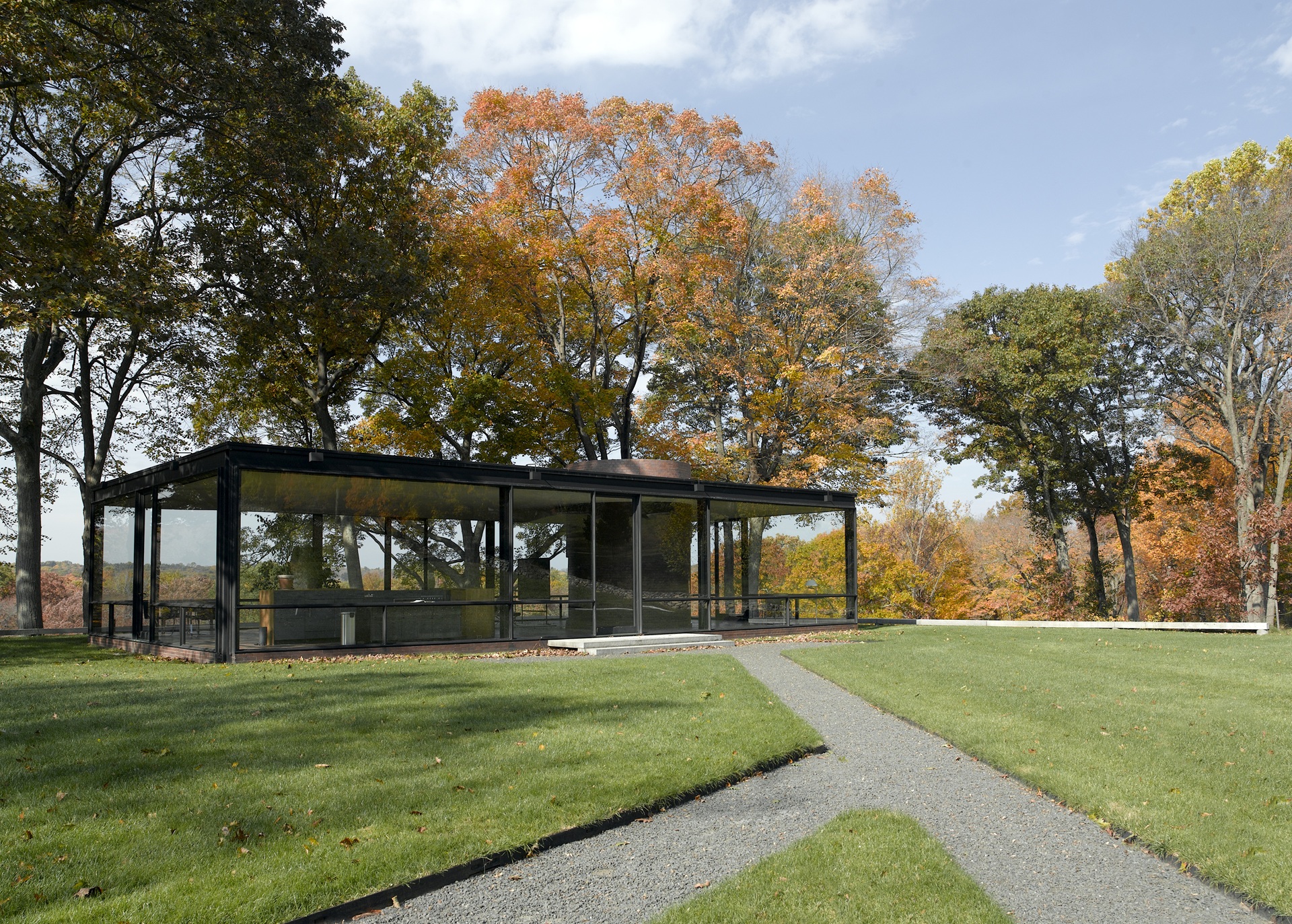 Vista da Glass House de Philip Johnson, com estrutura de aço escuro e paredes de vidro, cercada por gramado bem cuidado e árvores tingidas pelos tons alaranjados e dourados do outono em New Canaan, Connecticut