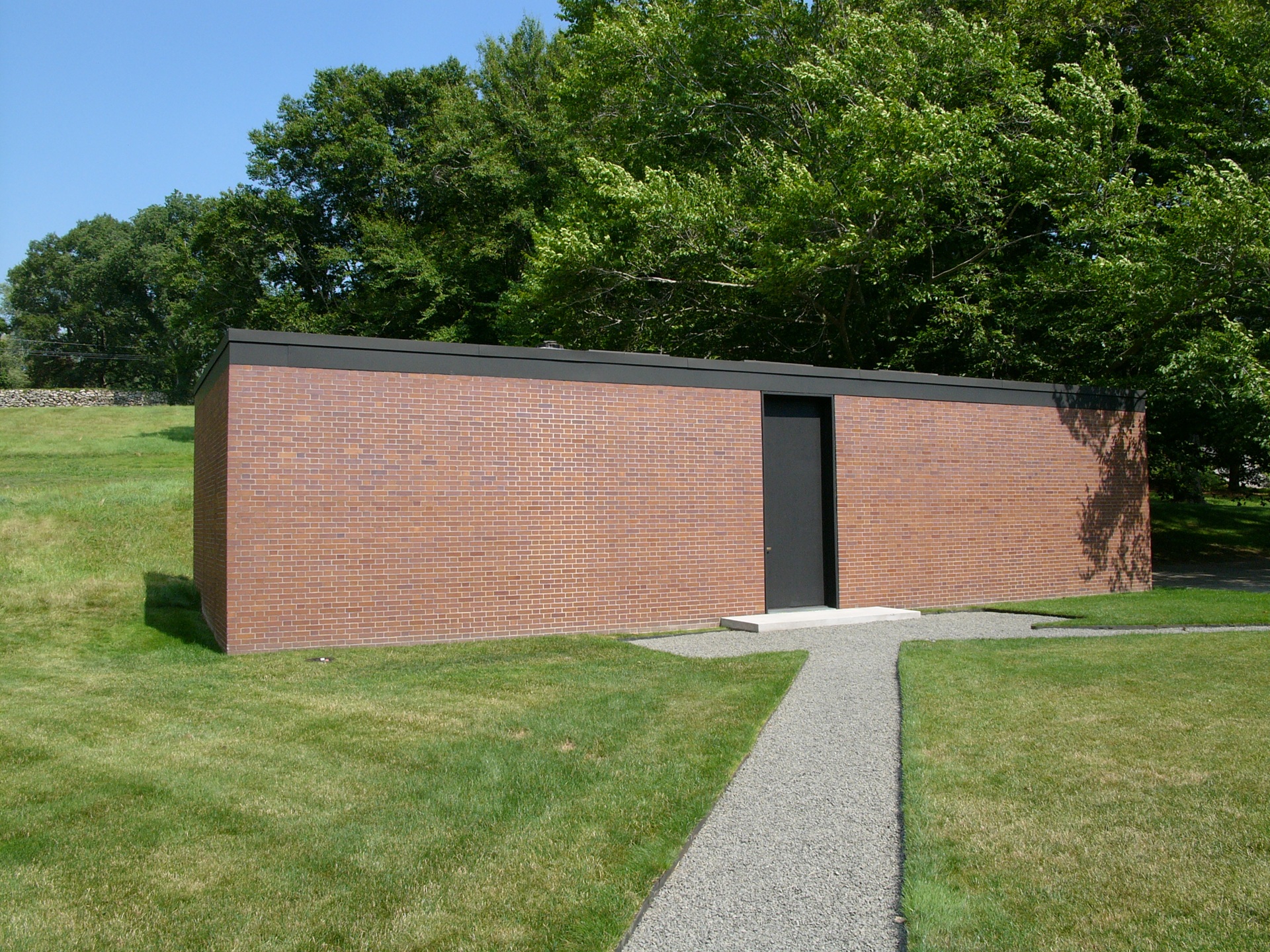 Fachada simples da Brick House, construção retangular de tijolos com porta preta central, rodeada por gramado e árvores na propriedade da Glass House