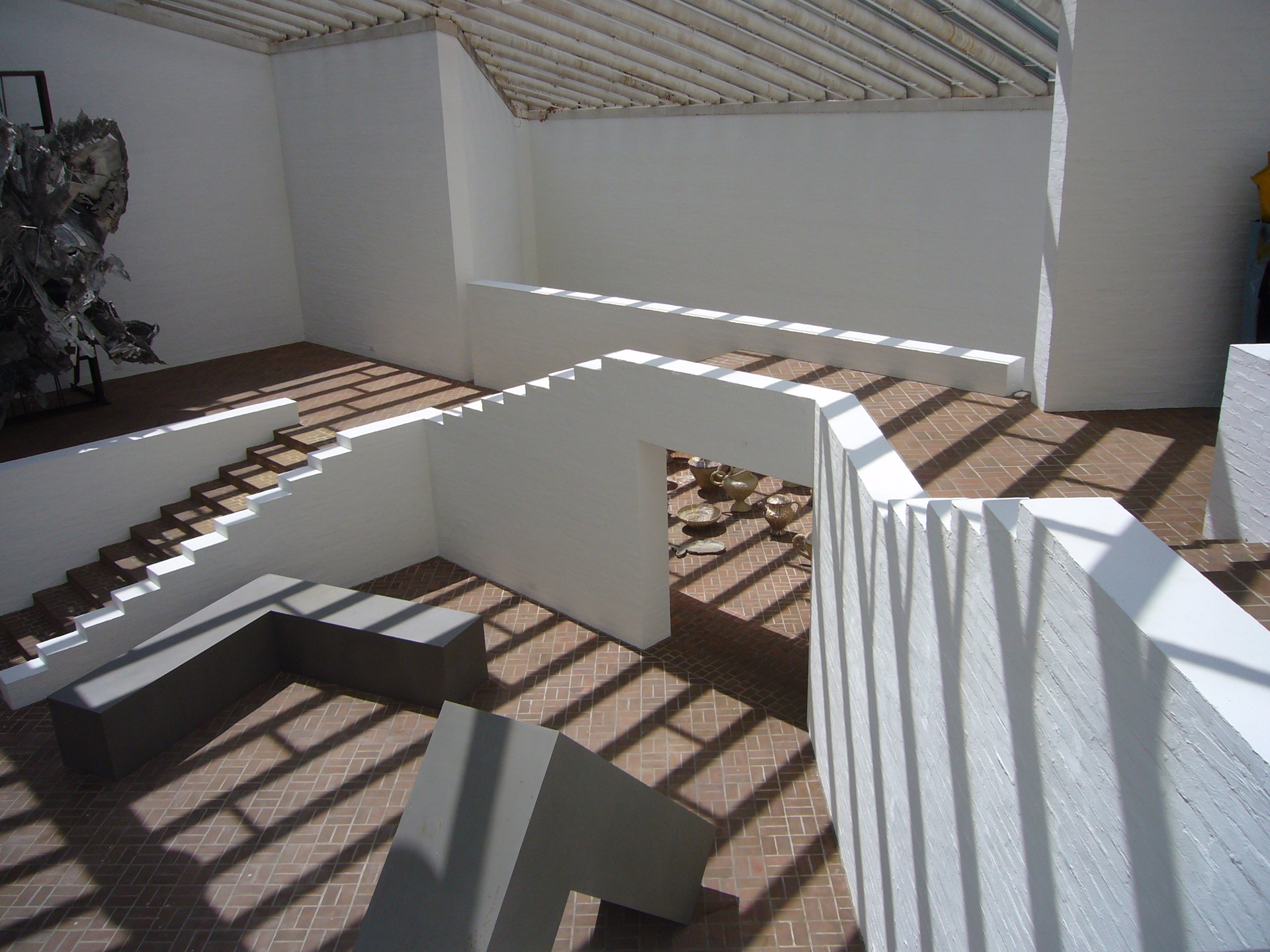 Interior da Galeria de Esculturas da Glass House, com paredes brancas, escadas geométricas, piso de tijolos e claraboias que projetam sombras marcantes sobre o espaço