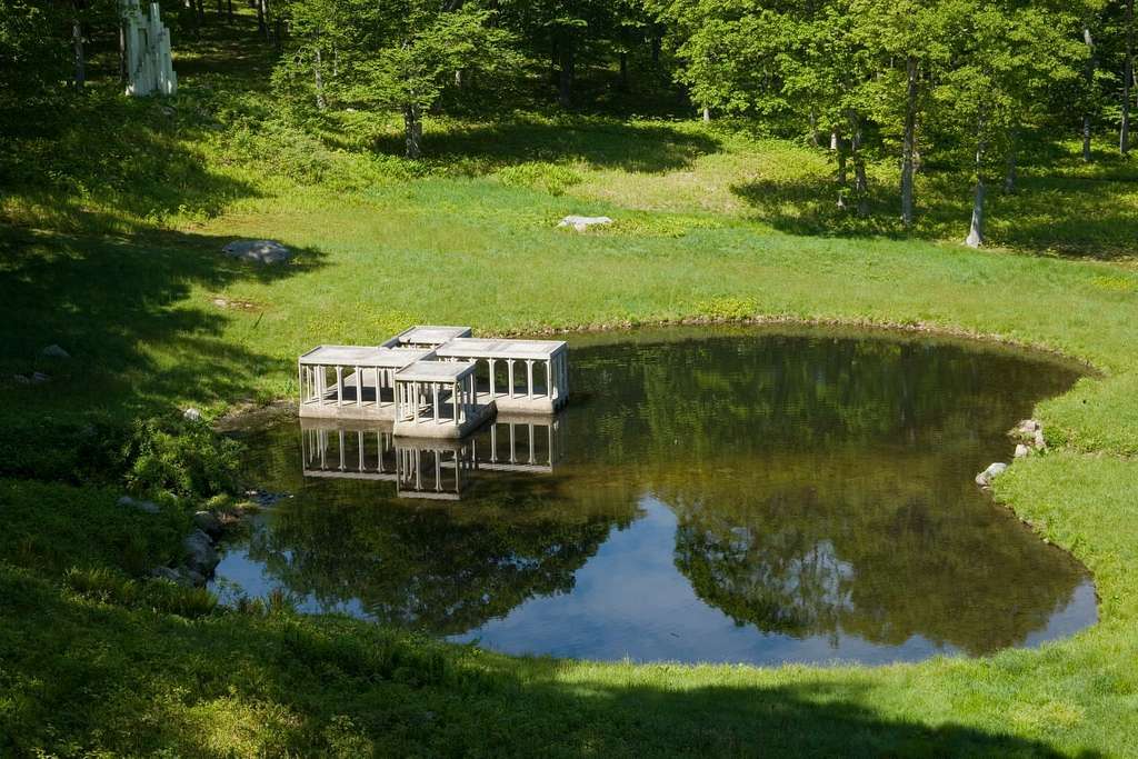 Vista aérea do Pavilhão no Lago, estrutura branca com colunas refletida na superfície de um pequeno lago cercado por vegetação e árvores