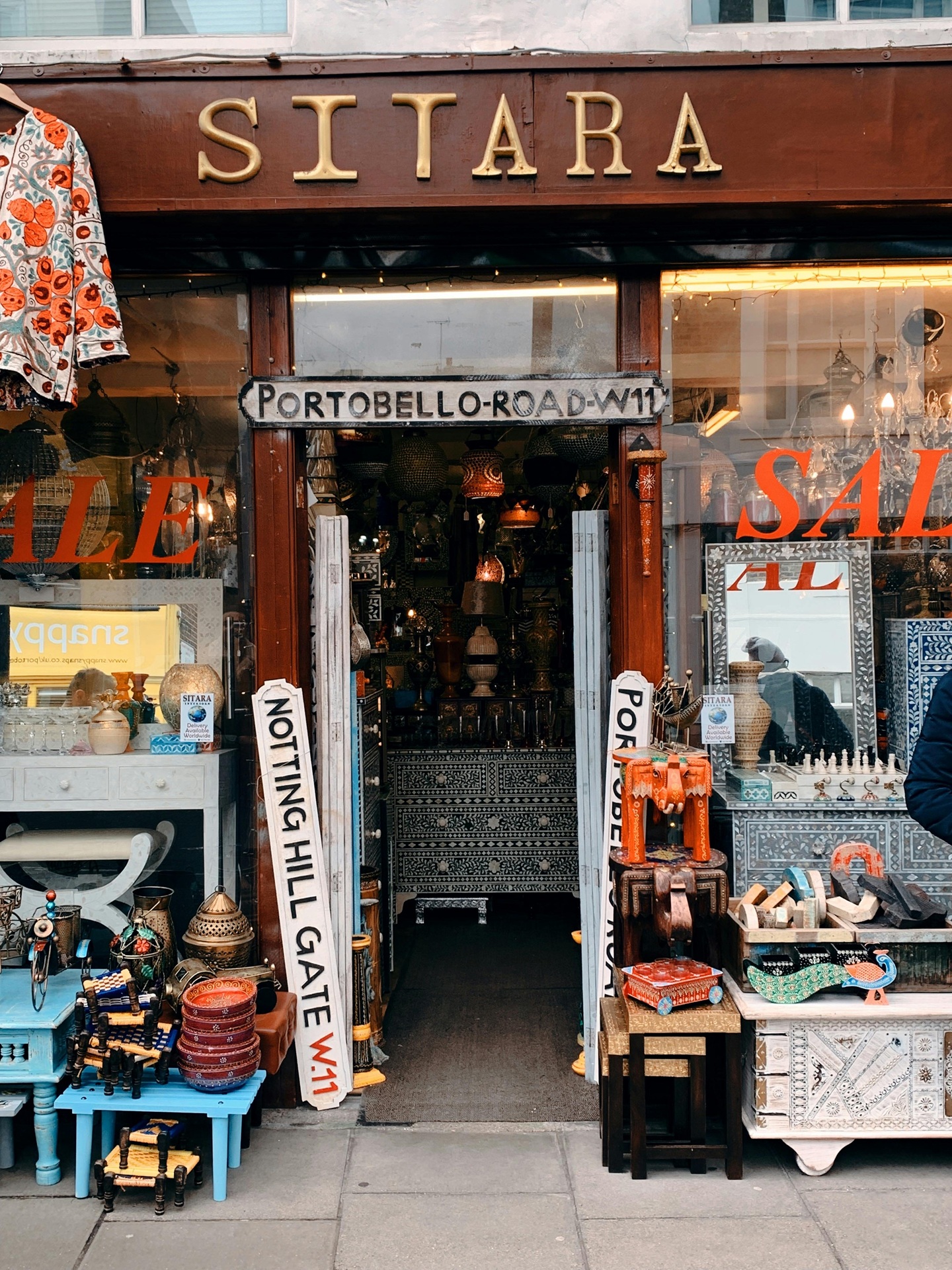 Fachada da loja Sitara, no Portobello Road Market, exibe objetos artesanais, móveis pintados e placas decorativas. O interior revela um ambiente repleto de luminárias e peças étnicas