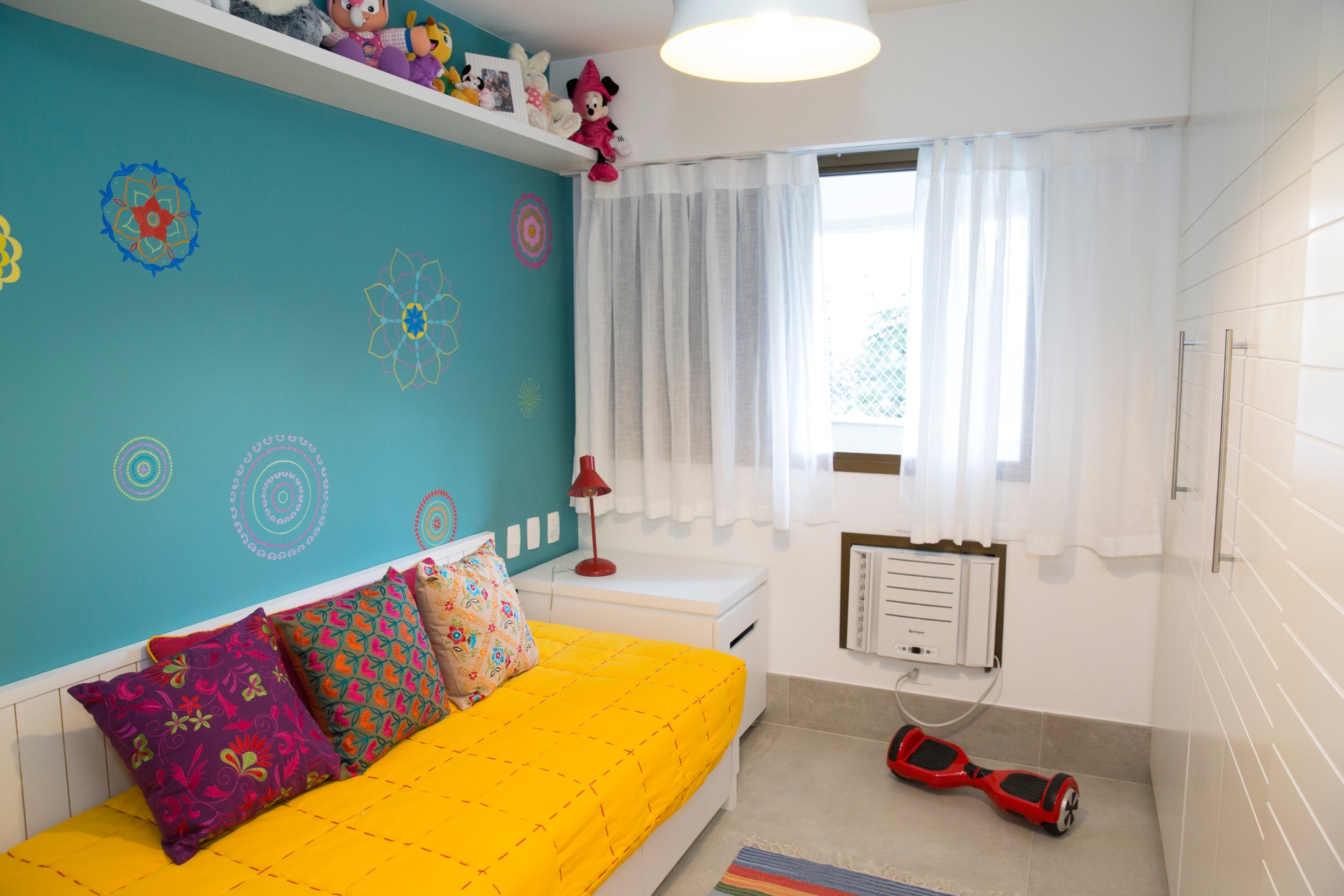 Quarto colorido em tons de amarelo, verde e rosa. Uma cama aparece à esquerda, enquanto na direita tem o guarda-roupa branco. Um hover board vermelho está no piso