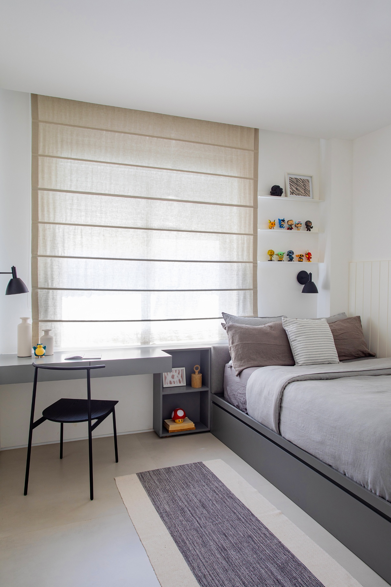 Quarto em tons neutros. Há uma cama à direita e uma escrivaninha cinza à esquerda