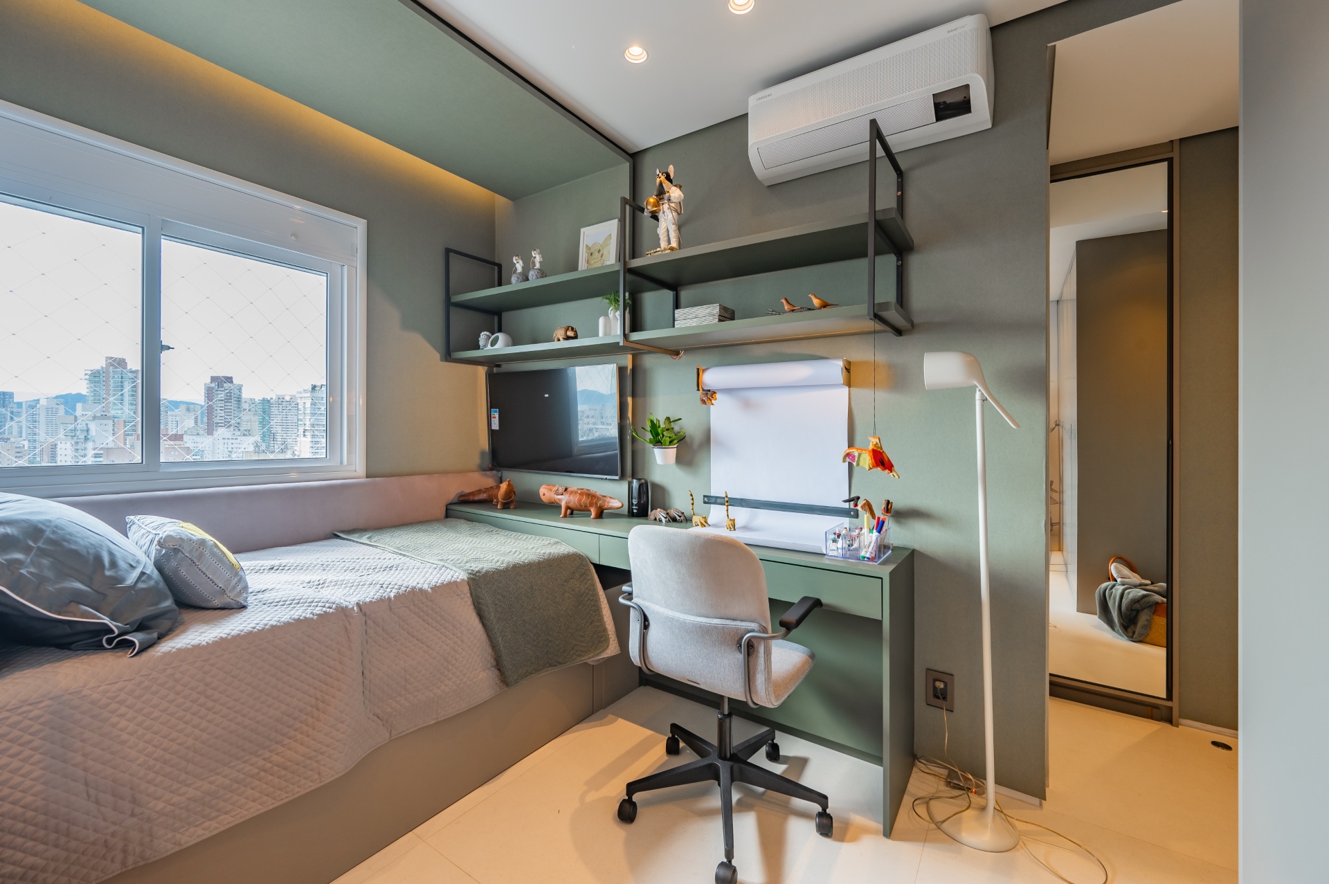Quarto com predominância de tons verdes. A escrivaninha se estende até a cama. Na parede temos uma prateleira e uma TV