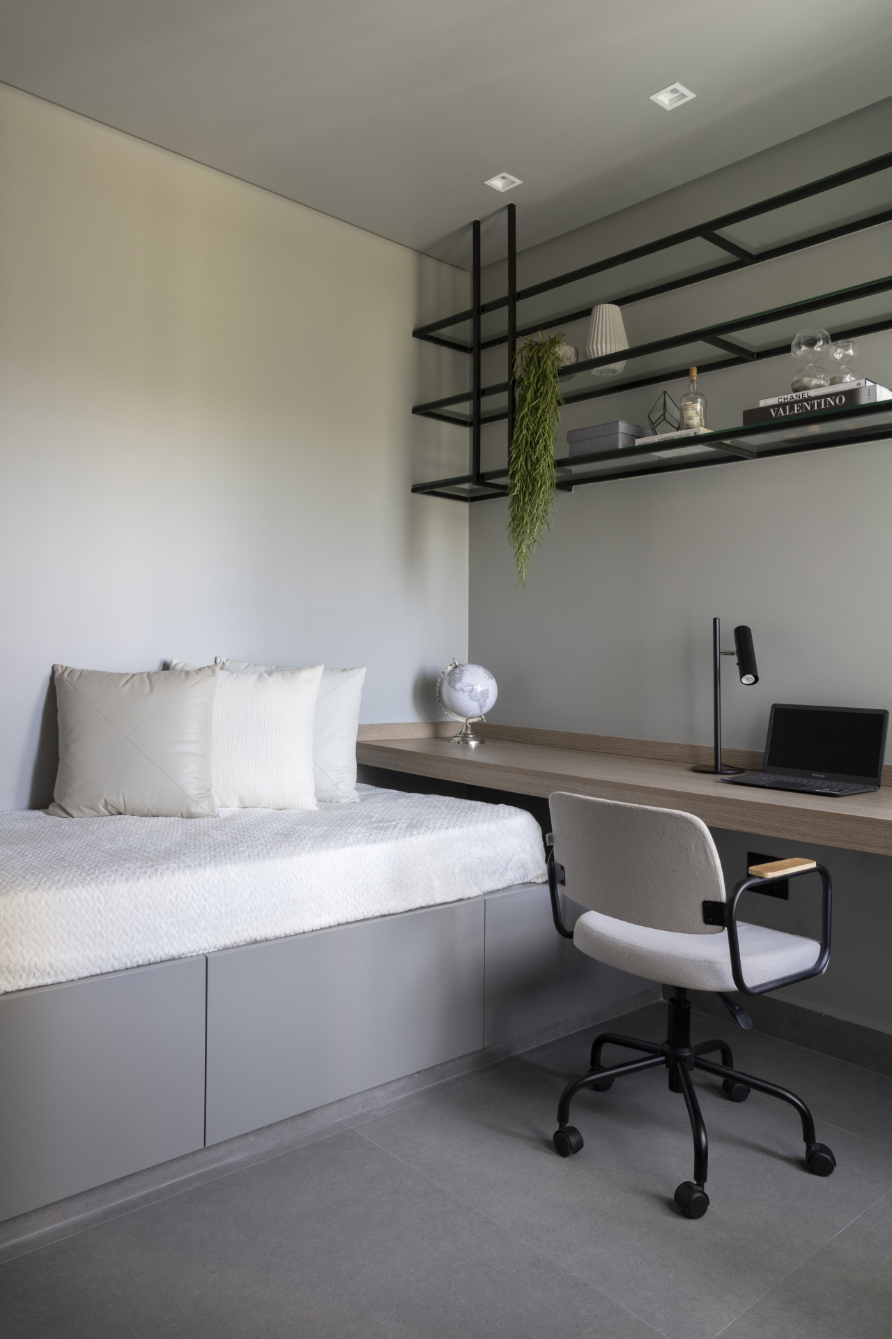 Quarto em tons neutros com predominância de cinza. A bancada da escrivaninha se estende até a cama, dando um visual de continuidade no espaço. Acima há uma prateleira suspensa em estilo industrial
