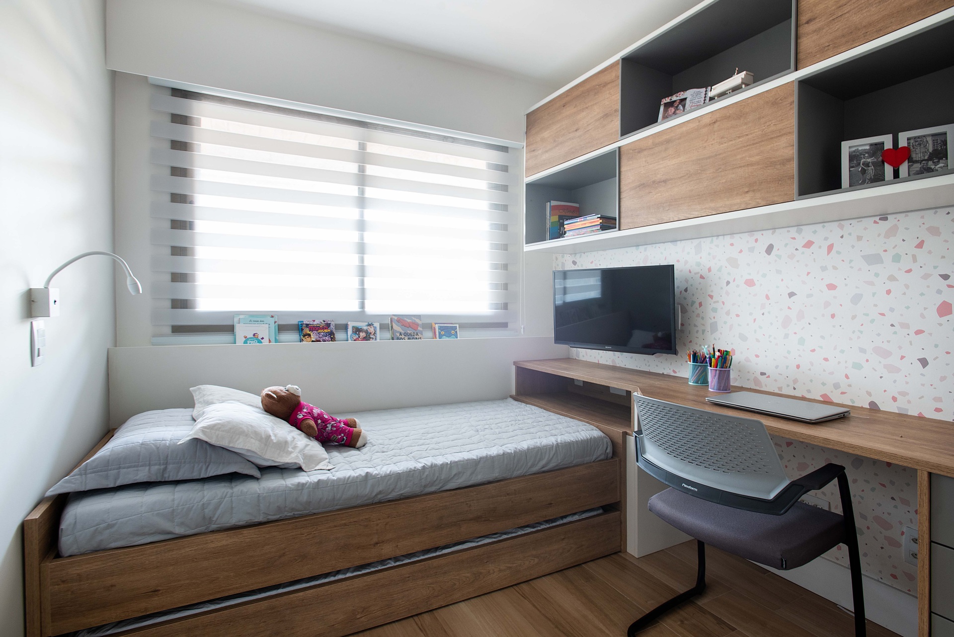 Quarto em tons neutros. À direita temos uma escrivaninha, nichos que pegam inteiramente a parede e uma TV. Ao fundo, encontra-se a cama abaixo da janela