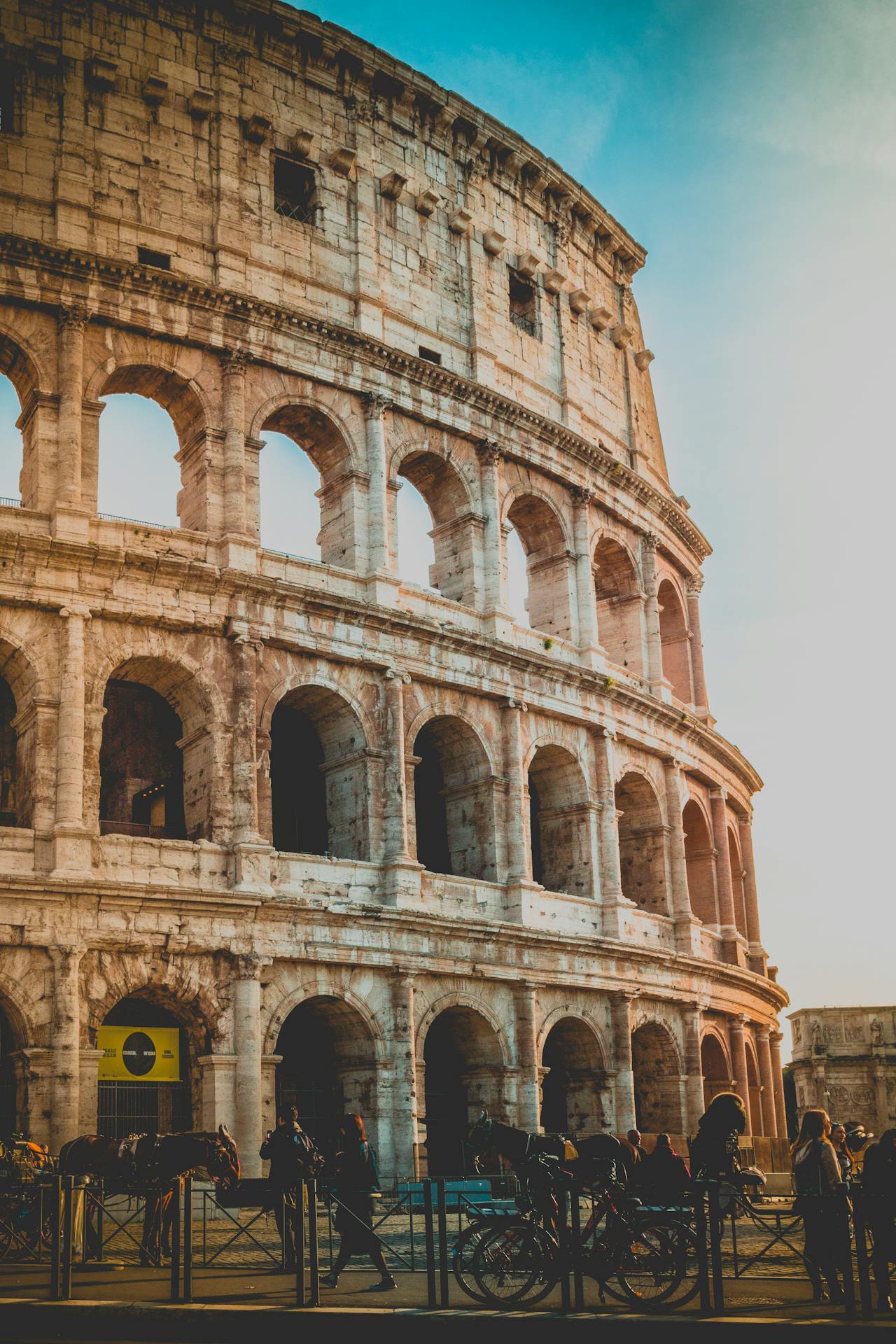 Fotografia do Coliseu em Roma, Itália, mostrando a sua estrutura de arcos e colunas em tom dourado pela luz do pôr do sol. Pessoas e bicicletas aparecem na parte inferior da imagem, destacando a interação entre o monumento histórico e a vida moderna