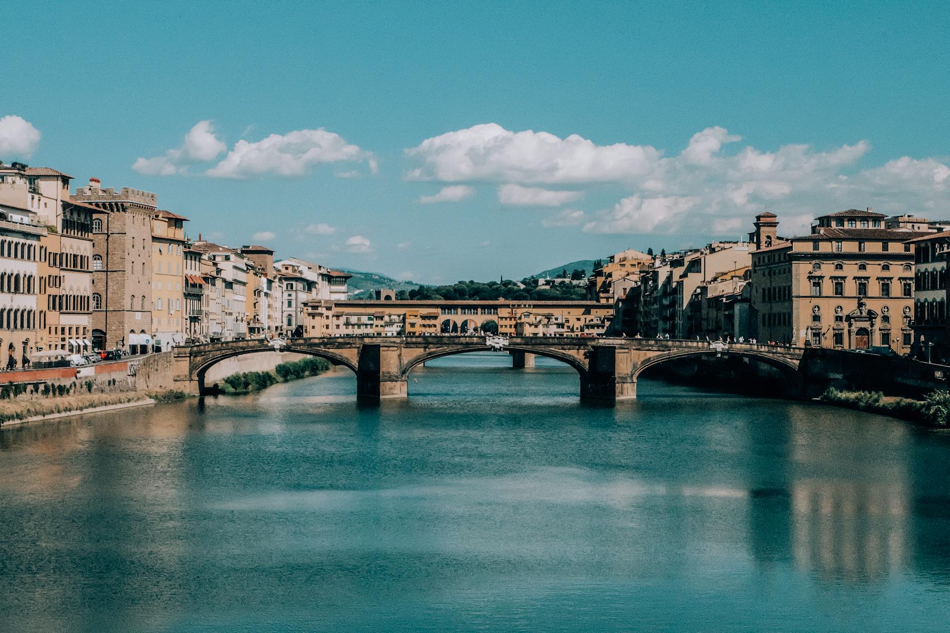 Vista da Ponte Vecchio, em Florença, Itália, com os seus arcos sustentando a estrutura sobre o Rio Arno. Edifícios históricos cercam a ponte sob um céu azul com algumas nuvens