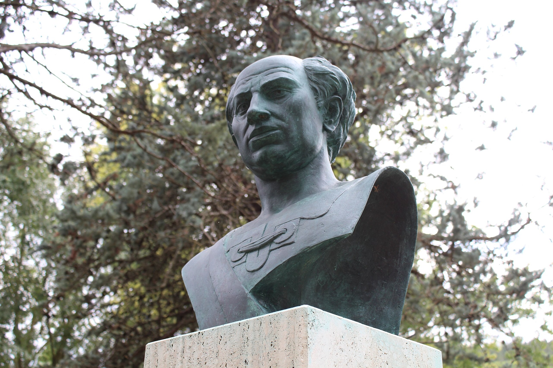 Busto de bronze de Donato Bramante sobre pedestal de pedra, localizado em área externa cercada por árvores. A escultura retrata o arquiteto renascentista com expressão séria e detalhes finos na vestimenta