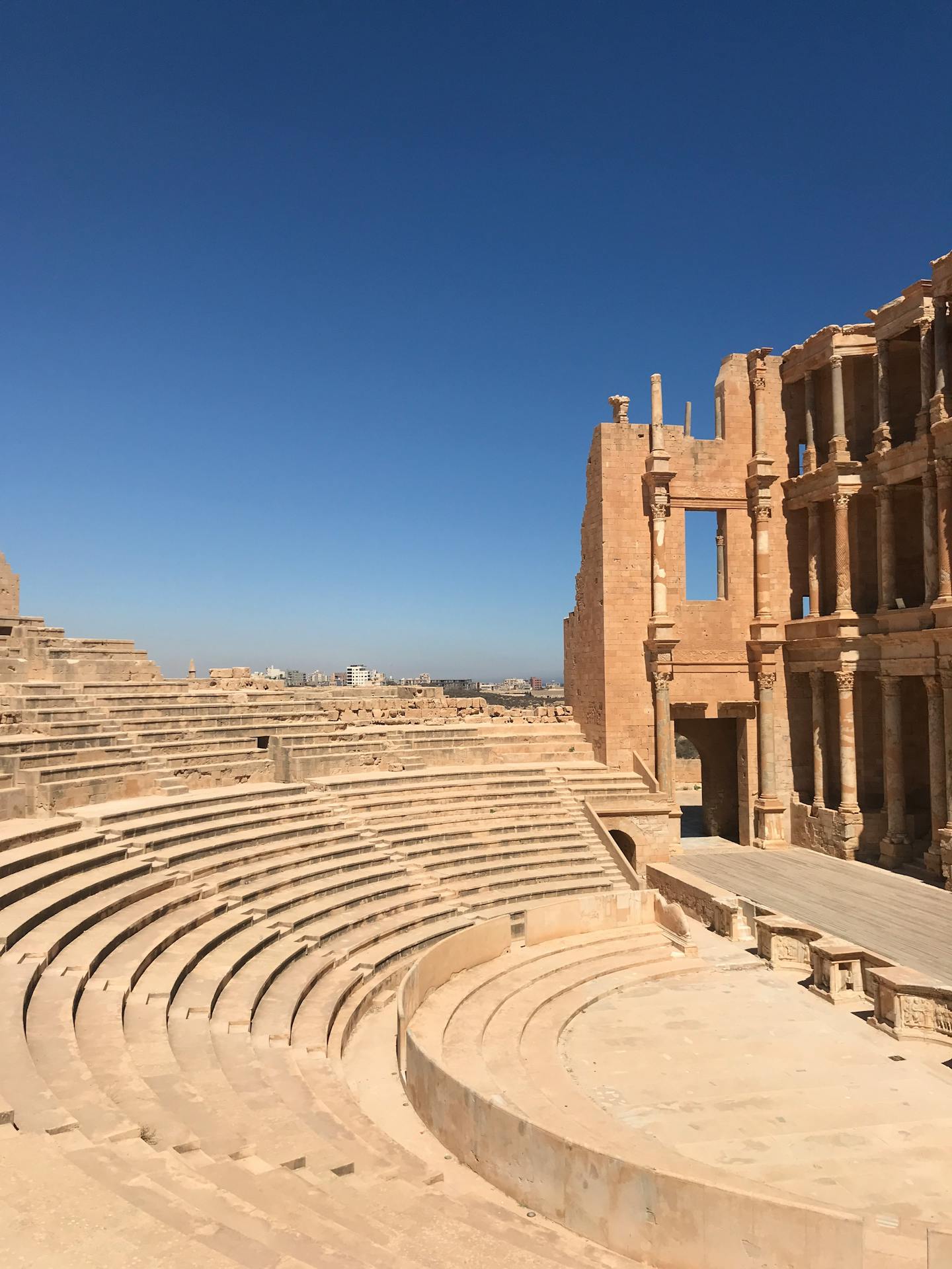 Vista de um anfiteatro romano antigo, com arquibancadas semicirculares de pedra e parte da estrutura ainda de pé, sob um céu azul intenso, destacando a arquitetura monumental clássica