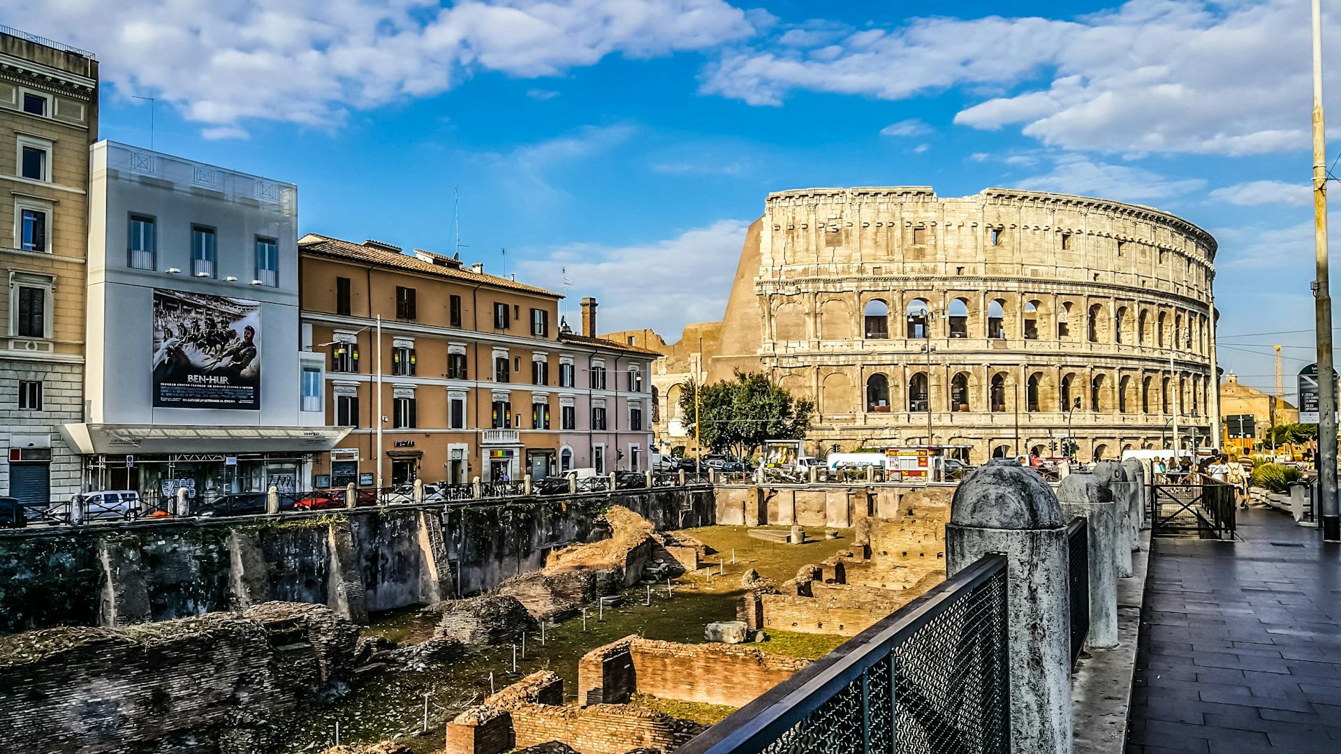 Vista urbana de Roma, Itália, com destaque para o Coliseu ao fundo. No primeiro plano, ruínas arqueológicas contrastam com edifícios modernos e ruas movimentadas sob um céu azul com nuvens