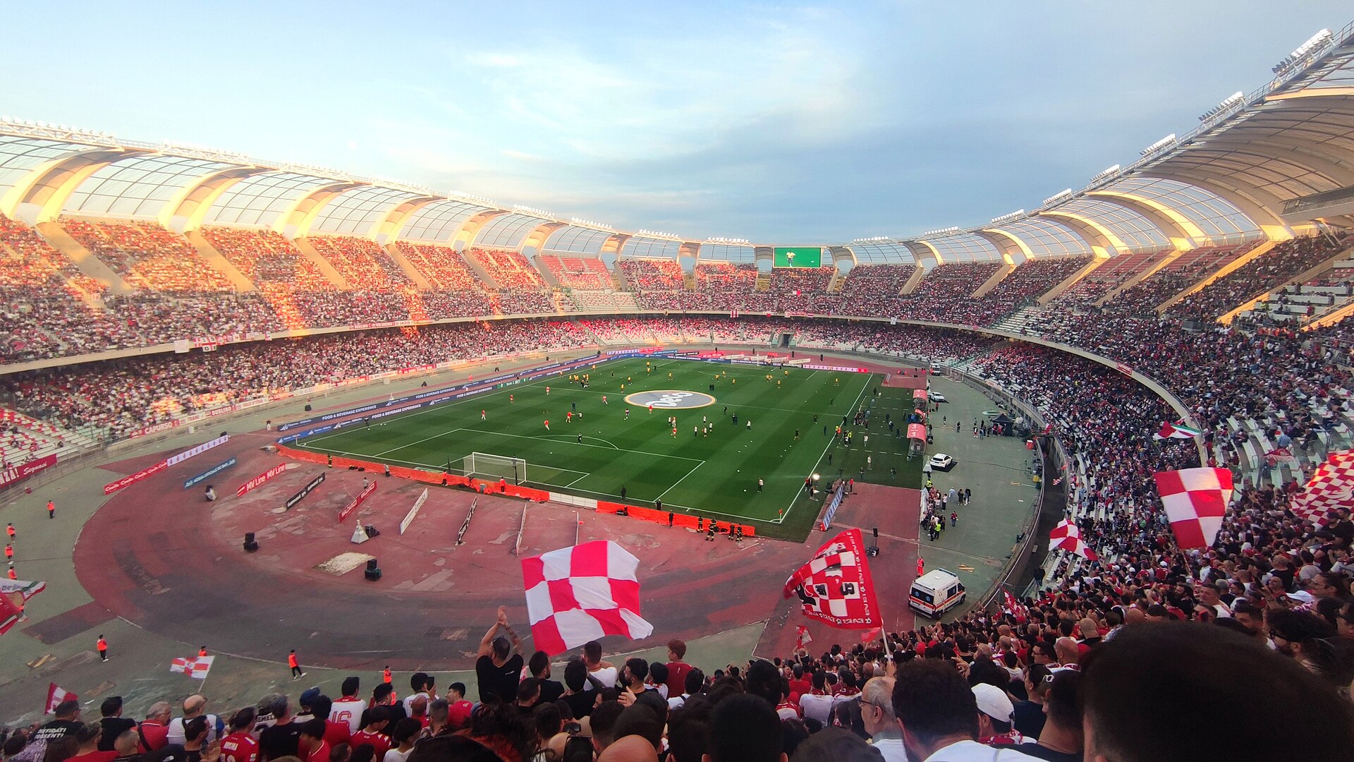 Estádio San Nicola, em Bari, Itália, repleto de torcedores durante uma partida de futebol. A estrutura apresenta arquibancadas curvas divididas em setores independentes, cercando o campo iluminado pelo pôr do sol