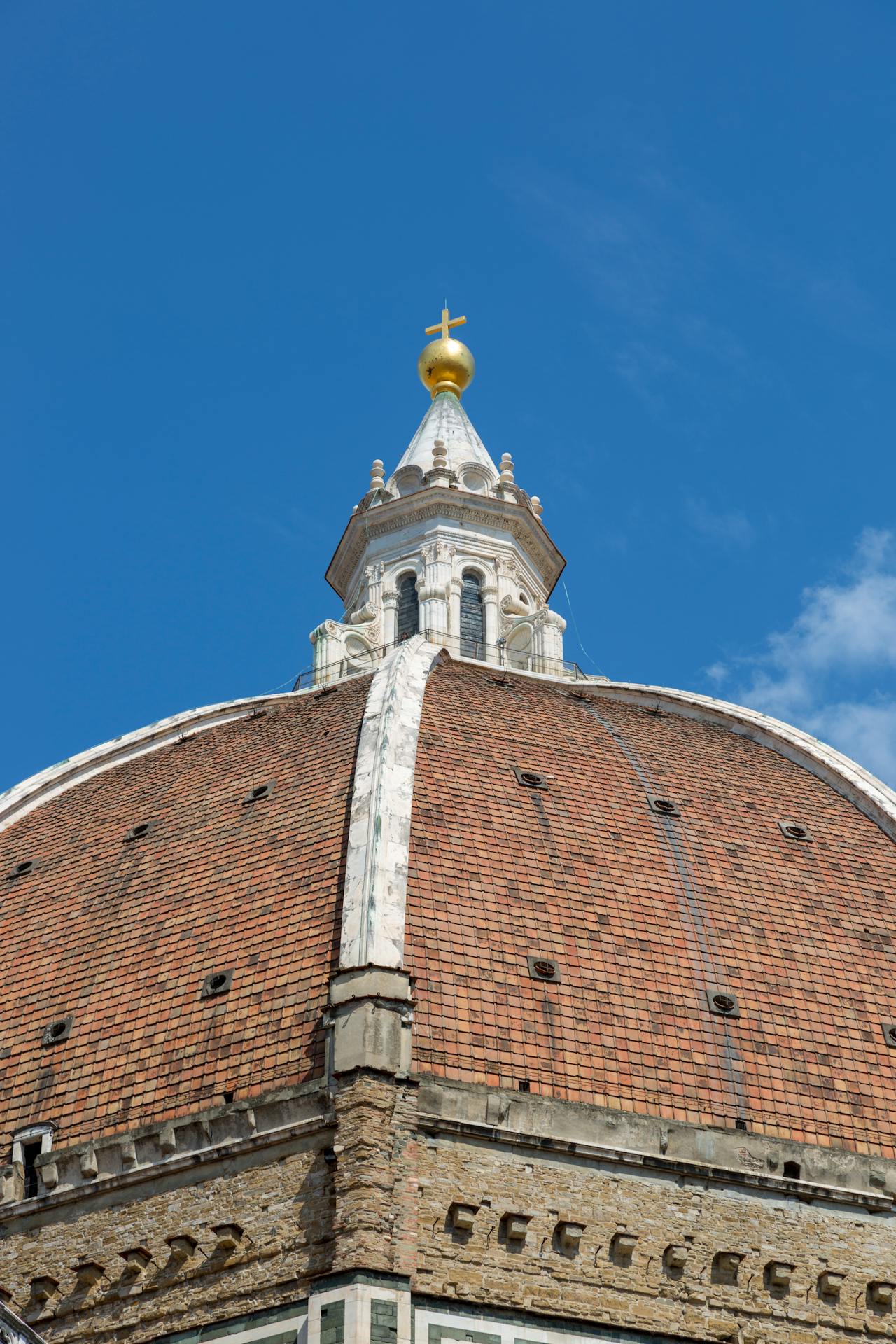 Detalhe da cúpula de Santa Maria del Fiore, em Florença, Itália, com revestimento em telhas vermelhas e topo branco decorado, finalizado por esfera dourada e cruz, sob um céu azul intenso