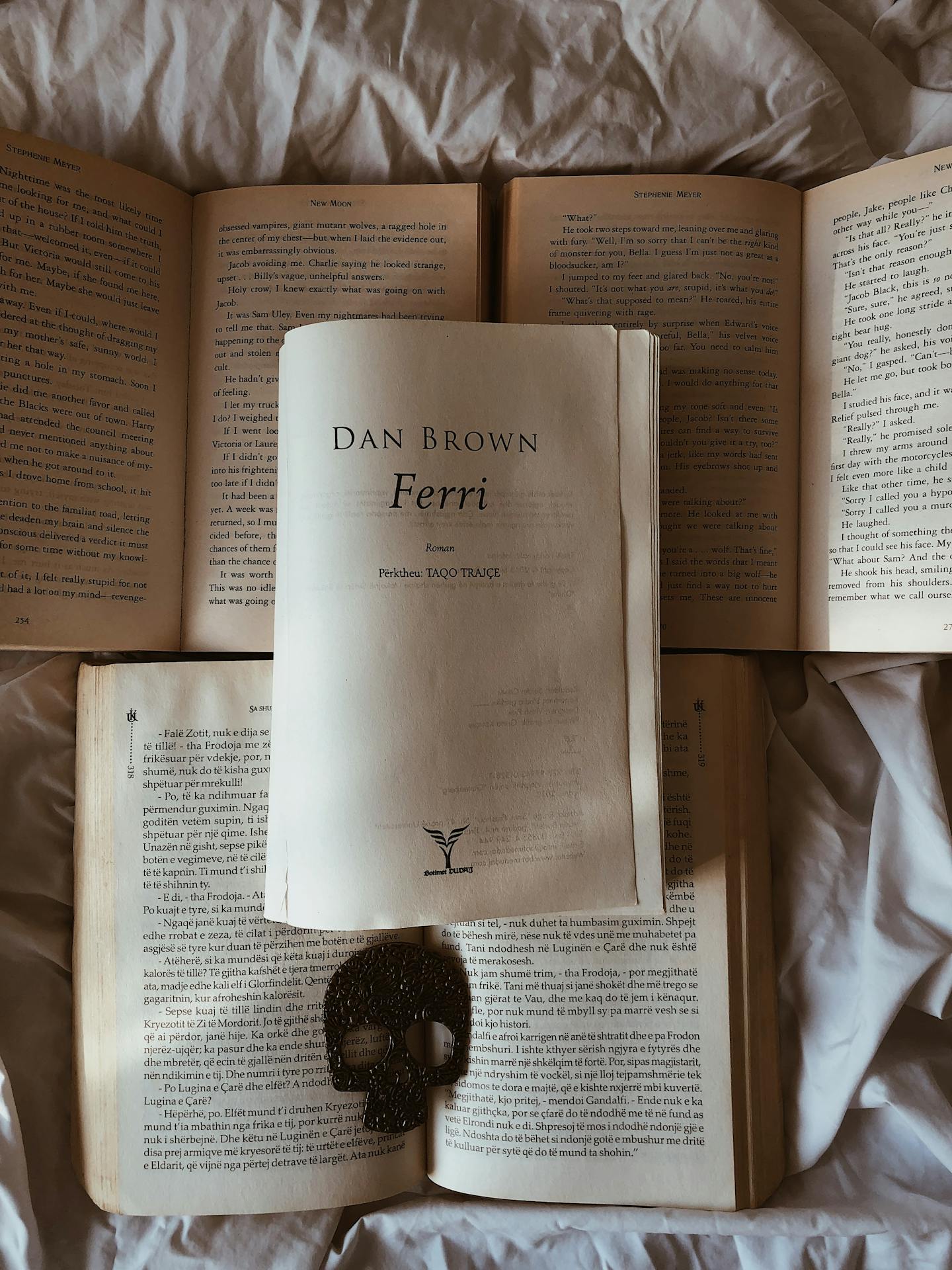 Livro de Dan Brown aberto sobre uma cama, cercado por outras páginas de livros, com um marcador em forma de caveira