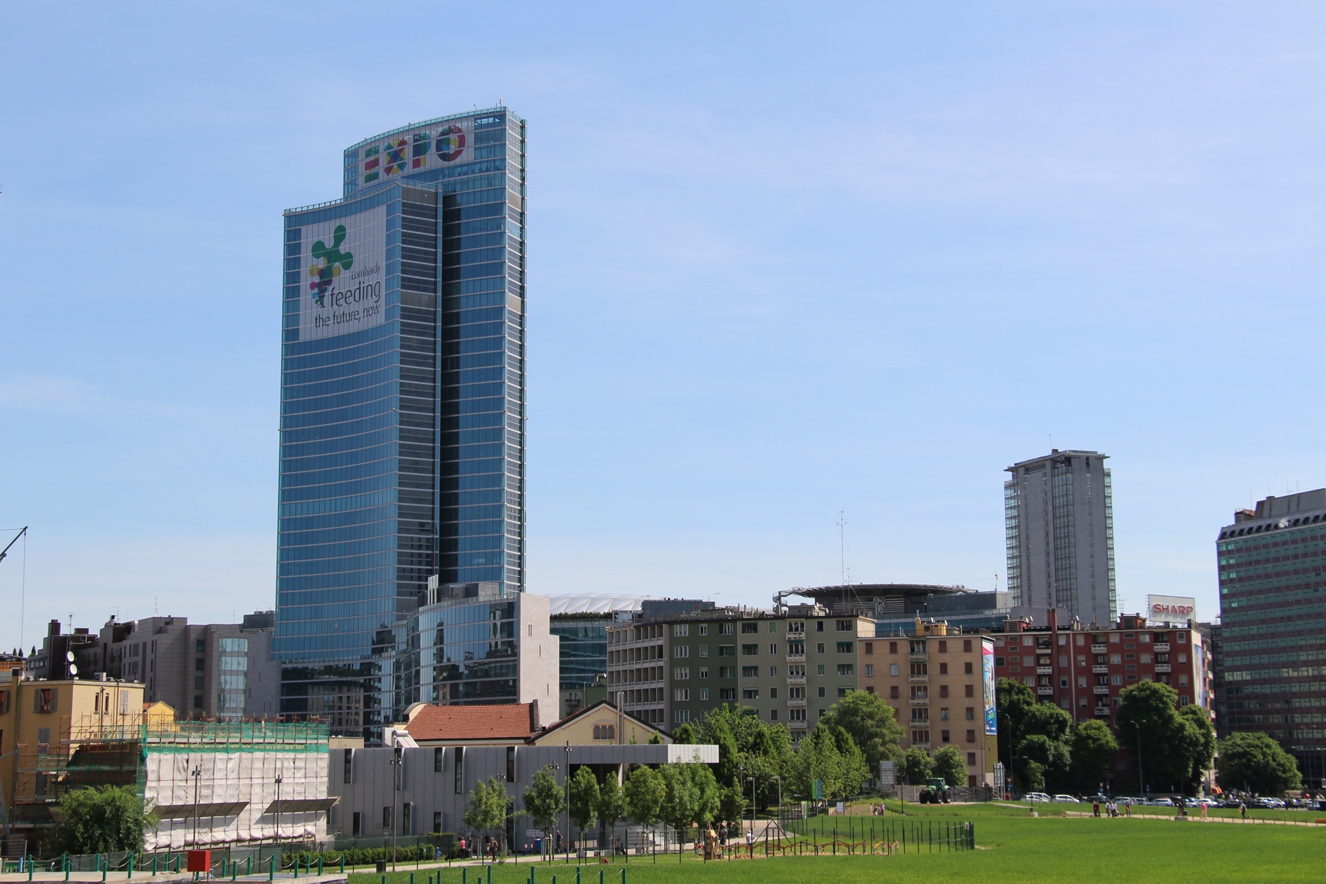 Vista do Palazzo Lombardia, em Milão, Itália, um arranha-céu moderno com fachada de vidro. Ao redor, edifícios mais baixos contrastam com a estrutura, simbolizando a integração entre passado e presente na paisagem urbana