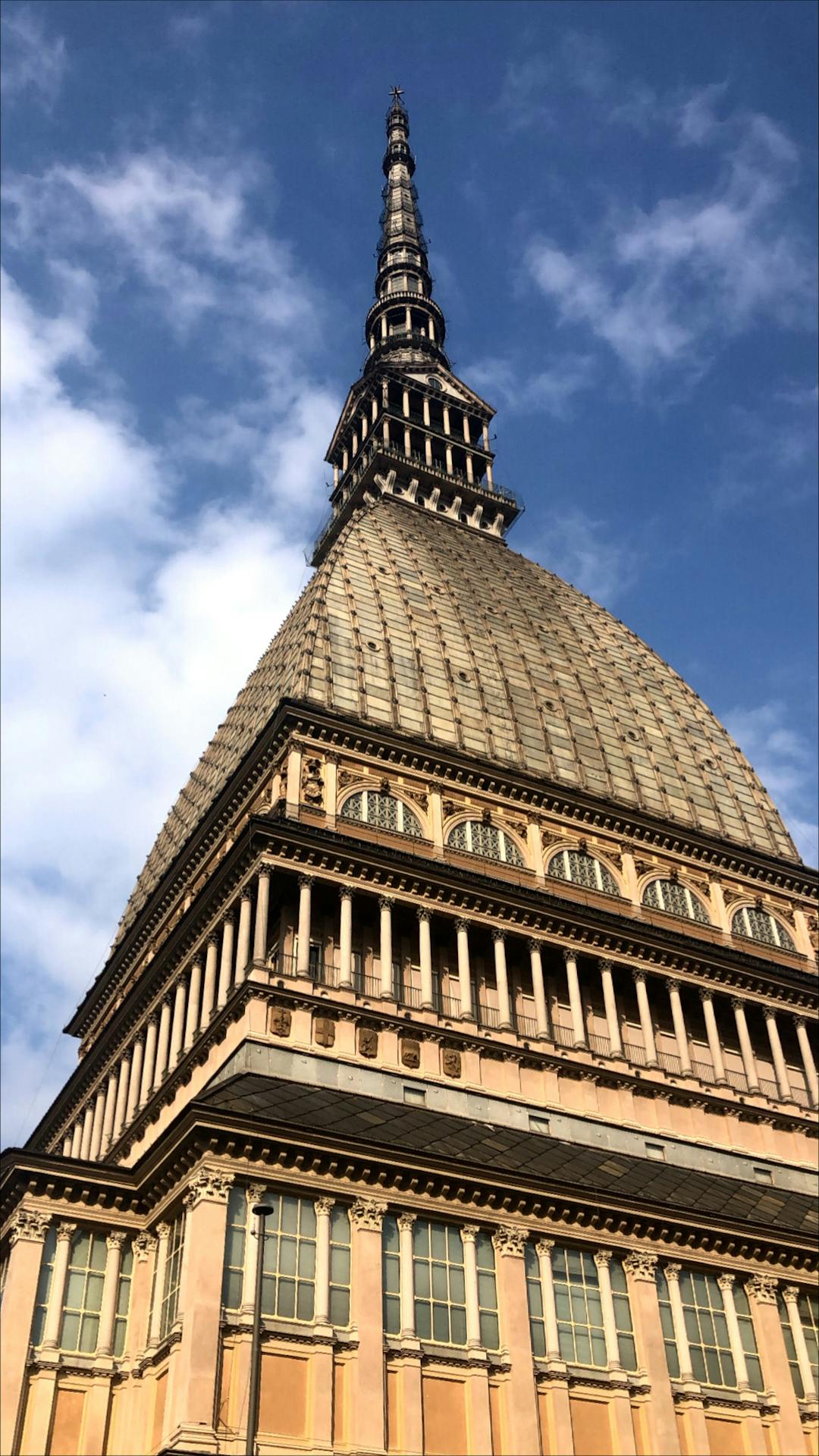 Vista em perspectiva da Mole Antonelliana, em Turim, Itália, com a sua cúpula imponente e torre elevada, sob um céu azul com nuvens esparsas