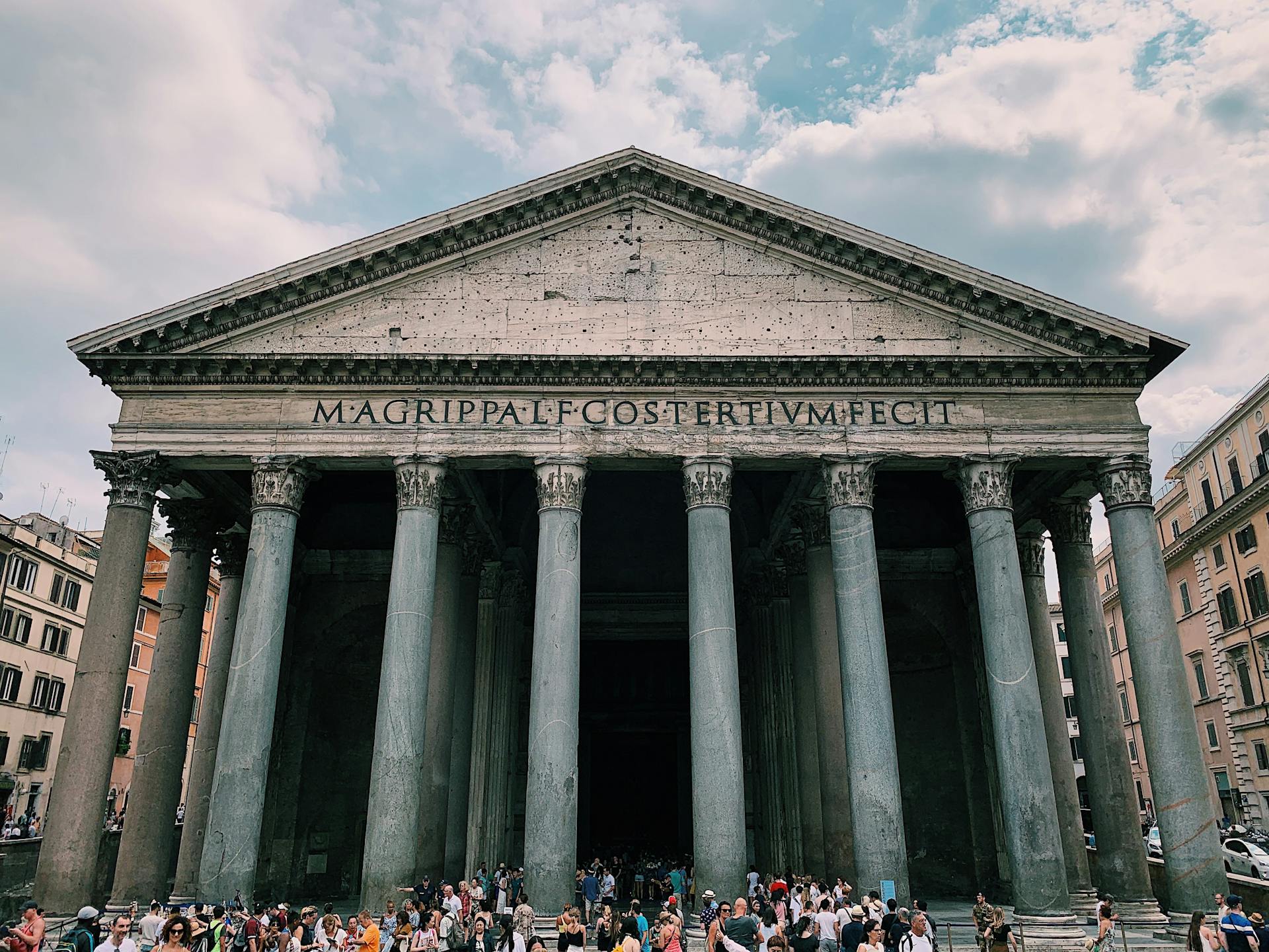 Fachada frontal do Panteão de Roma, com colunas coríntias maciças e inscrição em latim no frontão, cercado por turistas em uma praça movimentada