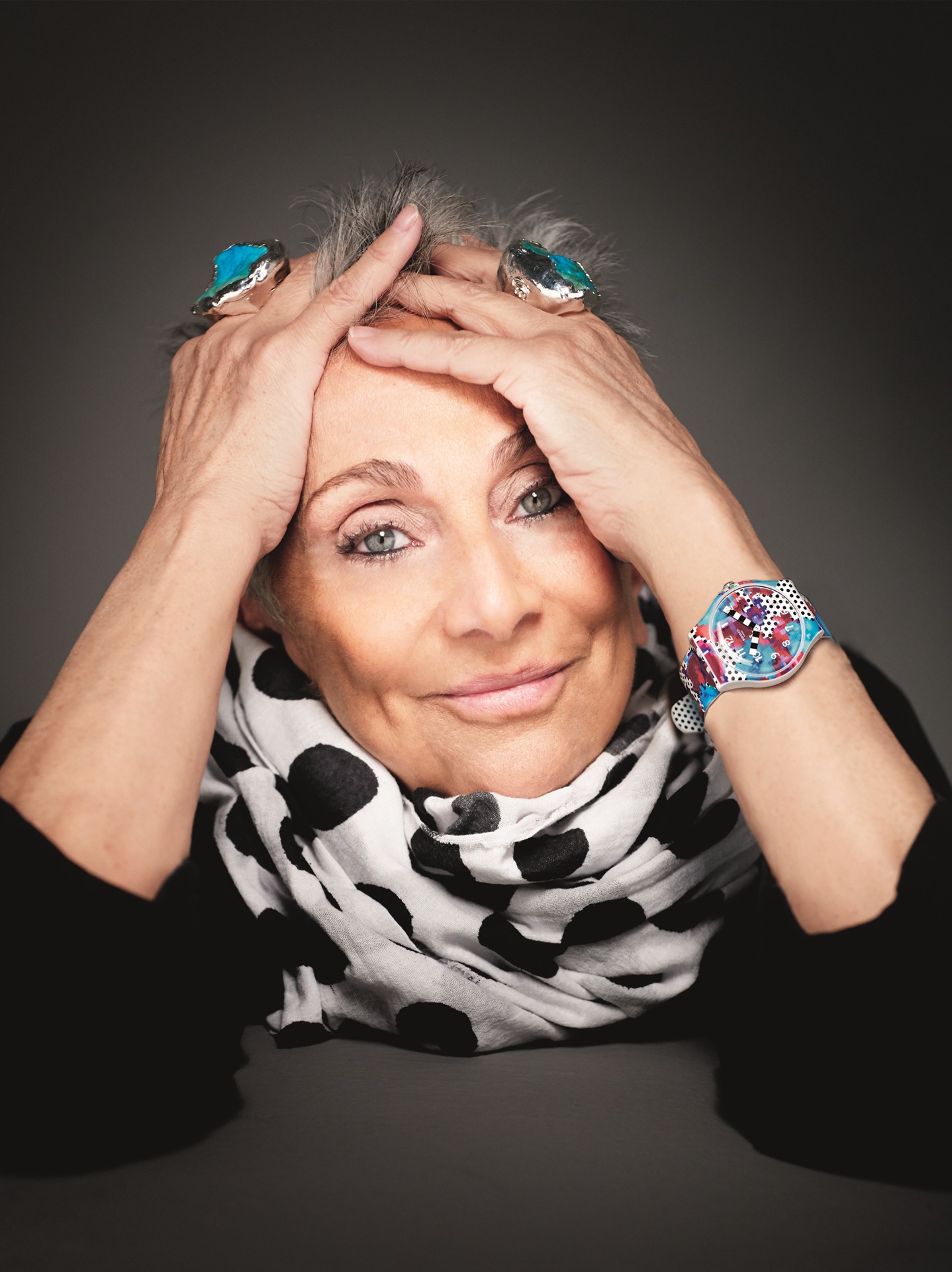Retrato de Paola Navone, sorridente, com as mãos apoiadas na cabeça. Ela usa anéis grandes com pedras azuis, relógio colorido e lenço branco com estampa de bolas pretas. Fundo neutro e iluminação suave destacam sua expressão criativa