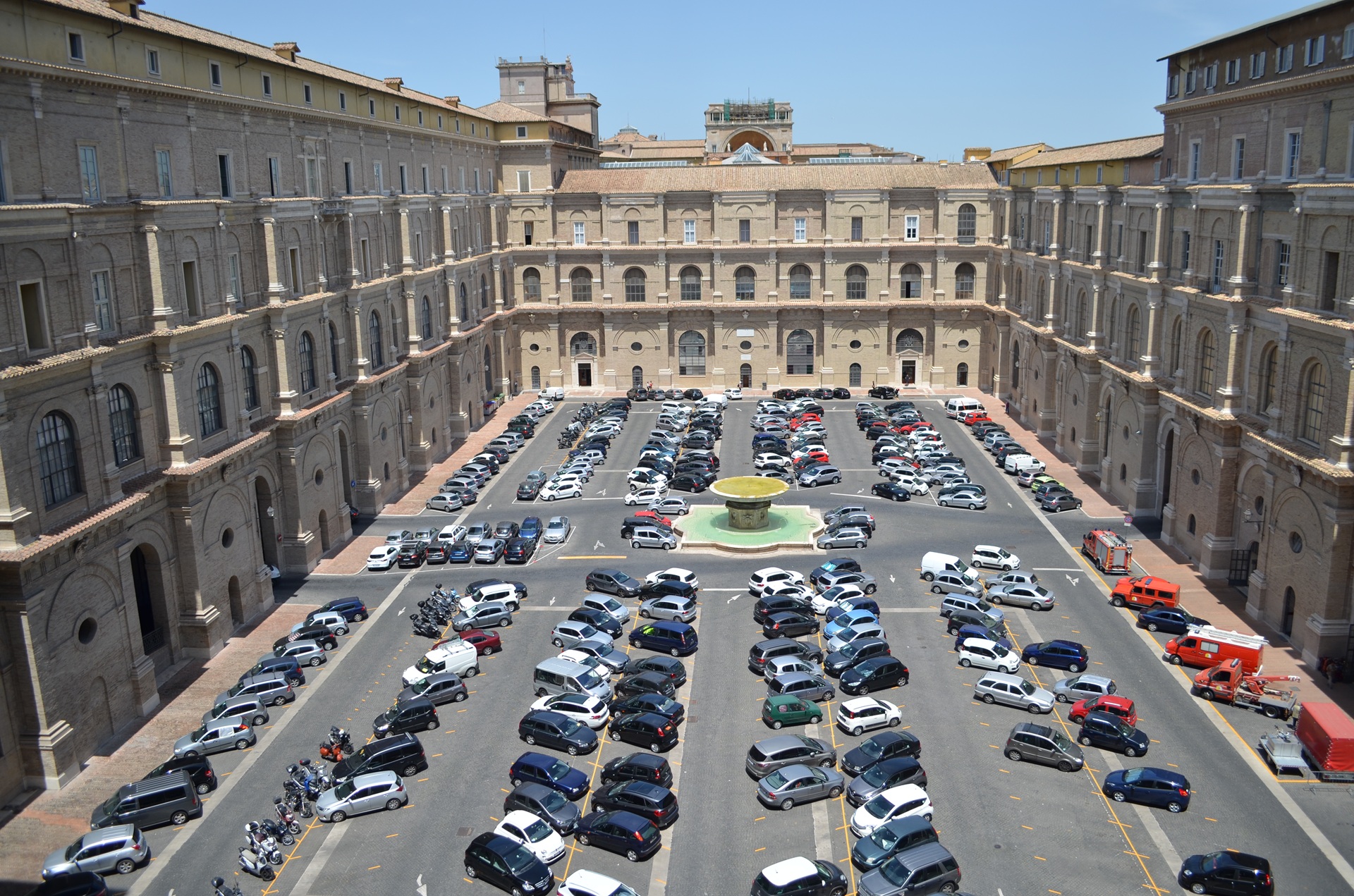 Vista interna de um pátio no Vaticano, cercado por edifícios de arquitetura clássica, com várias janelas e arcos. No centro, área aberta com carros estacionados, iluminada pela luz natural