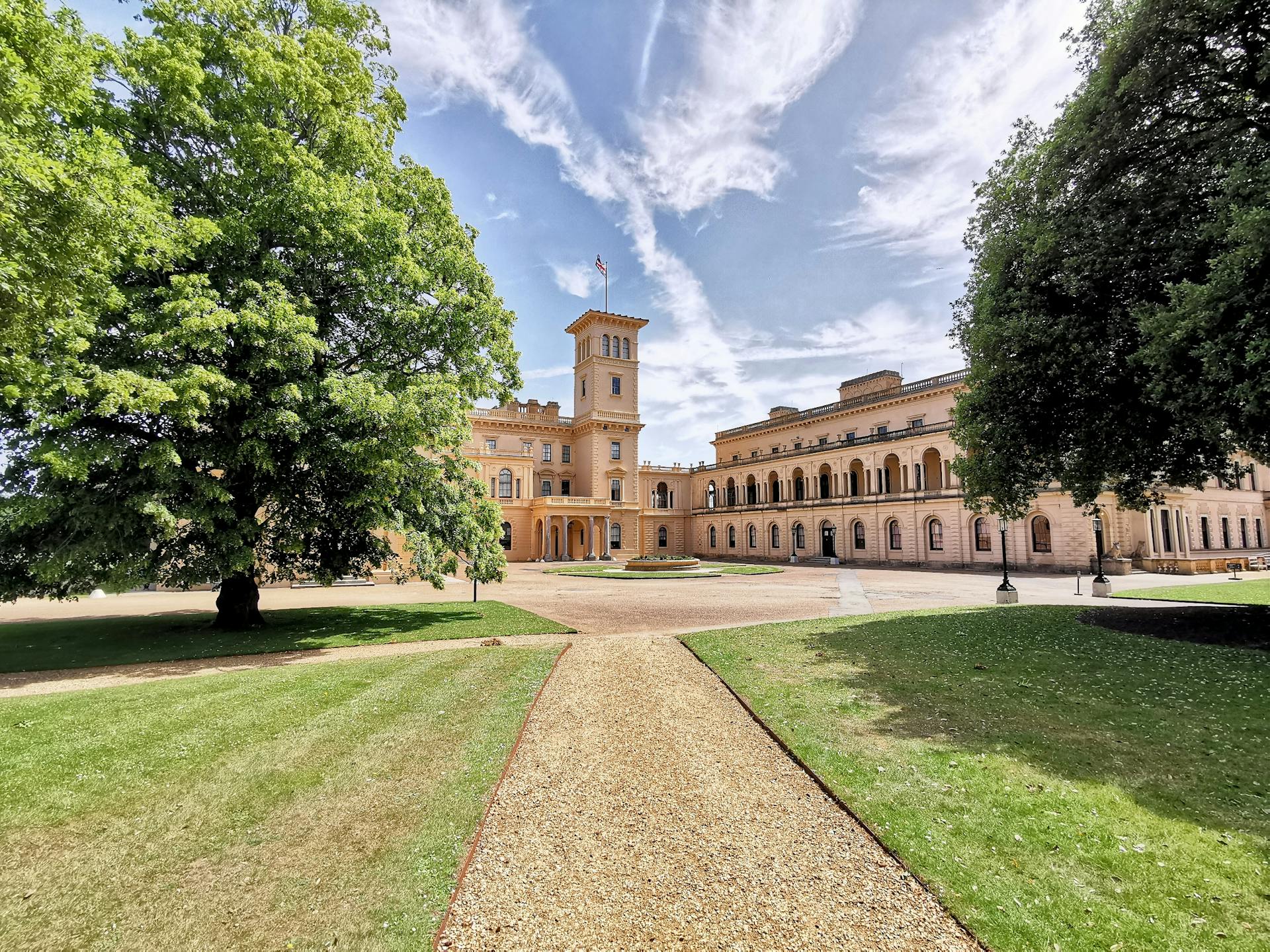 Vista frontal da Osborne House, na Ilha de Wight, Inglaterra. O edifício apresenta torre central, cornijas proeminentes e janelas em arco, rodeado por jardins verdes e árvores sob um céu azul com nuvens