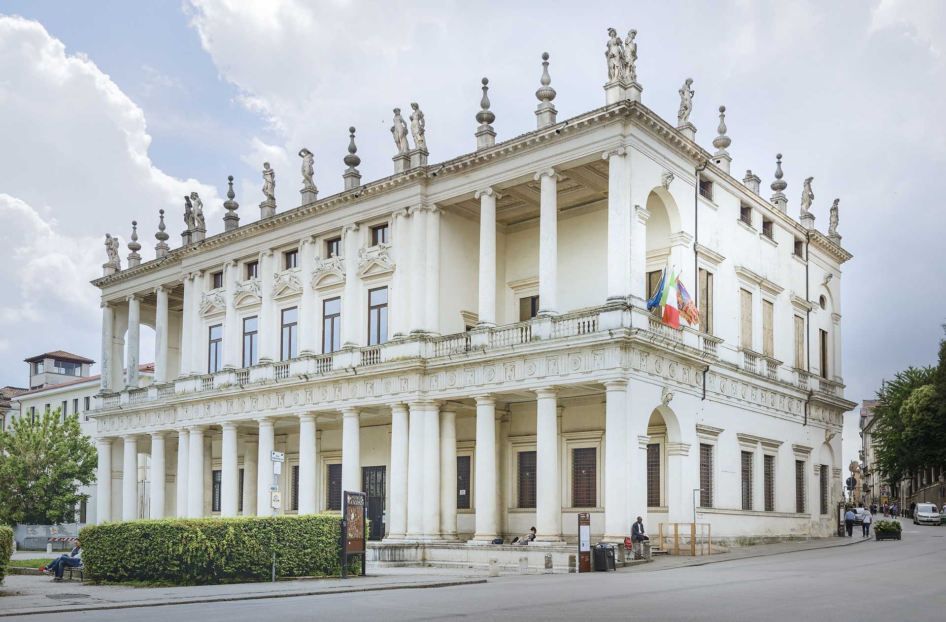 Imagem do Palazzo Chiericati, em Vicenza, Itália. O edifício renascentista apresenta colunas simétricas na fachada, esculturas no topo e bandeiras na entrada, com pessoas caminhando e sentadas ao redor