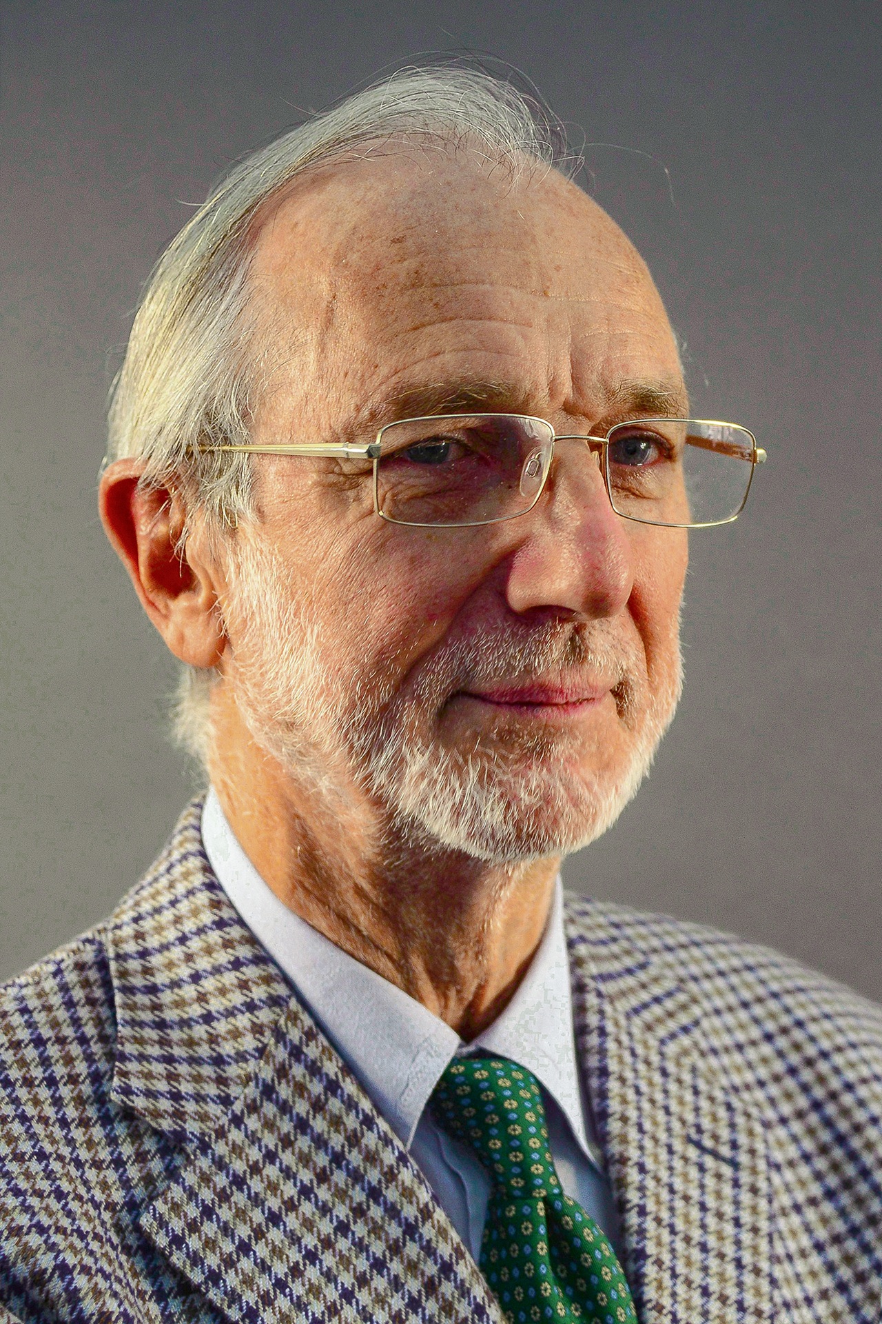 Retrato de Renzo Piano, arquiteto italiano, com expressão serena. Ele usa óculos, paletó com estampa xadrez, camisa clara e gravata verde estampada, em fundo neutro
