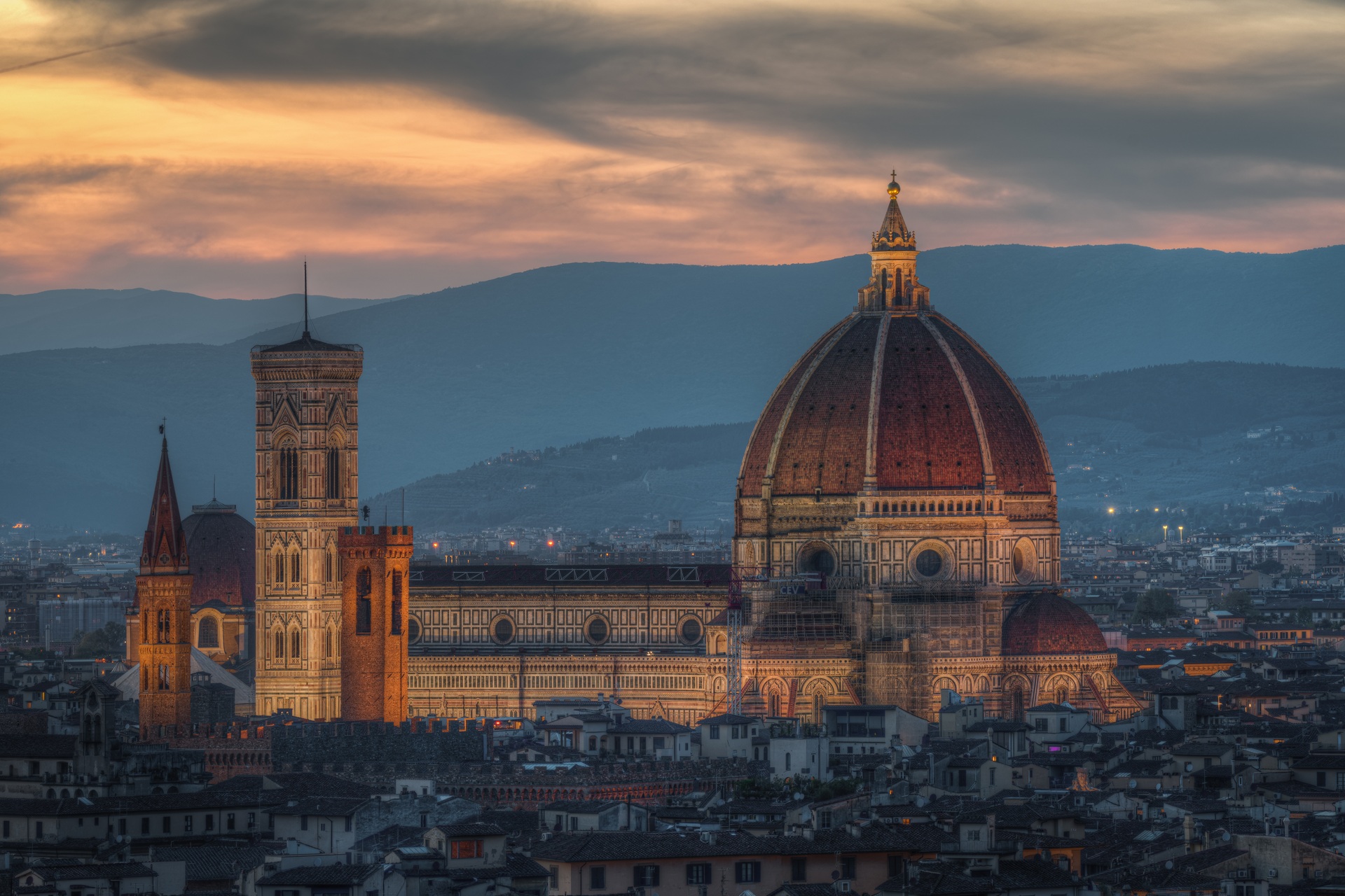 Vista panorâmica da Catedral de Santa Maria del Fiore, em Florença, Itália, com a sua imponente cúpula iluminada ao entardecer, cercada por construções históricas e montanhas ao fundo