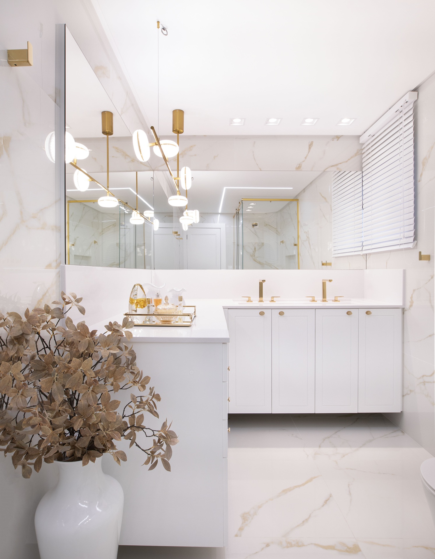 Banheiro com porcelanato marmorizado branco, gabinetes brancos e detalhes dourados