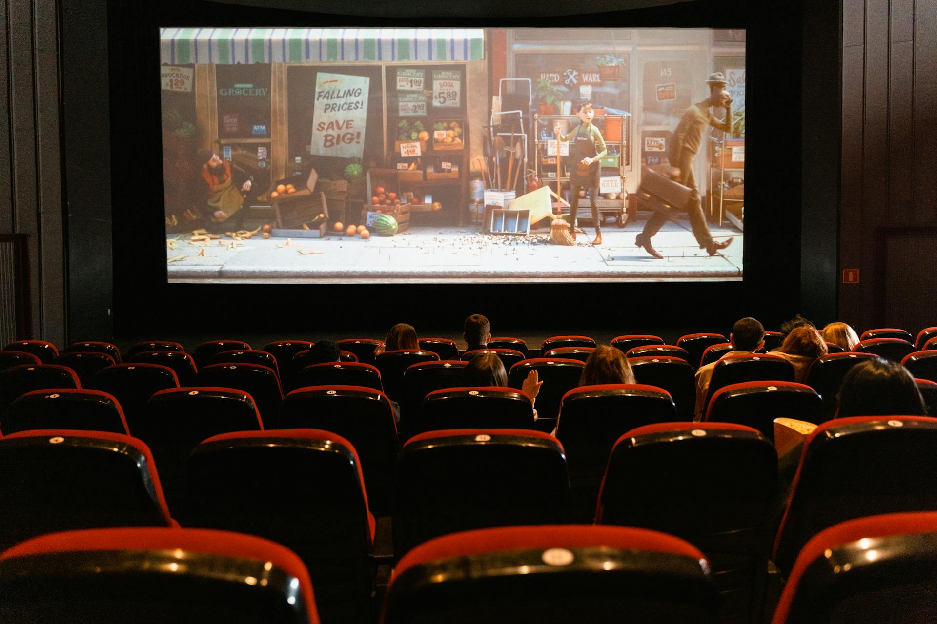 Visão interna de uma sala de cinema com poltronas vermelhas em primeiro plano e, ao fundo, a tela projeta uma cena animada enquanto alguns espectadores assistem